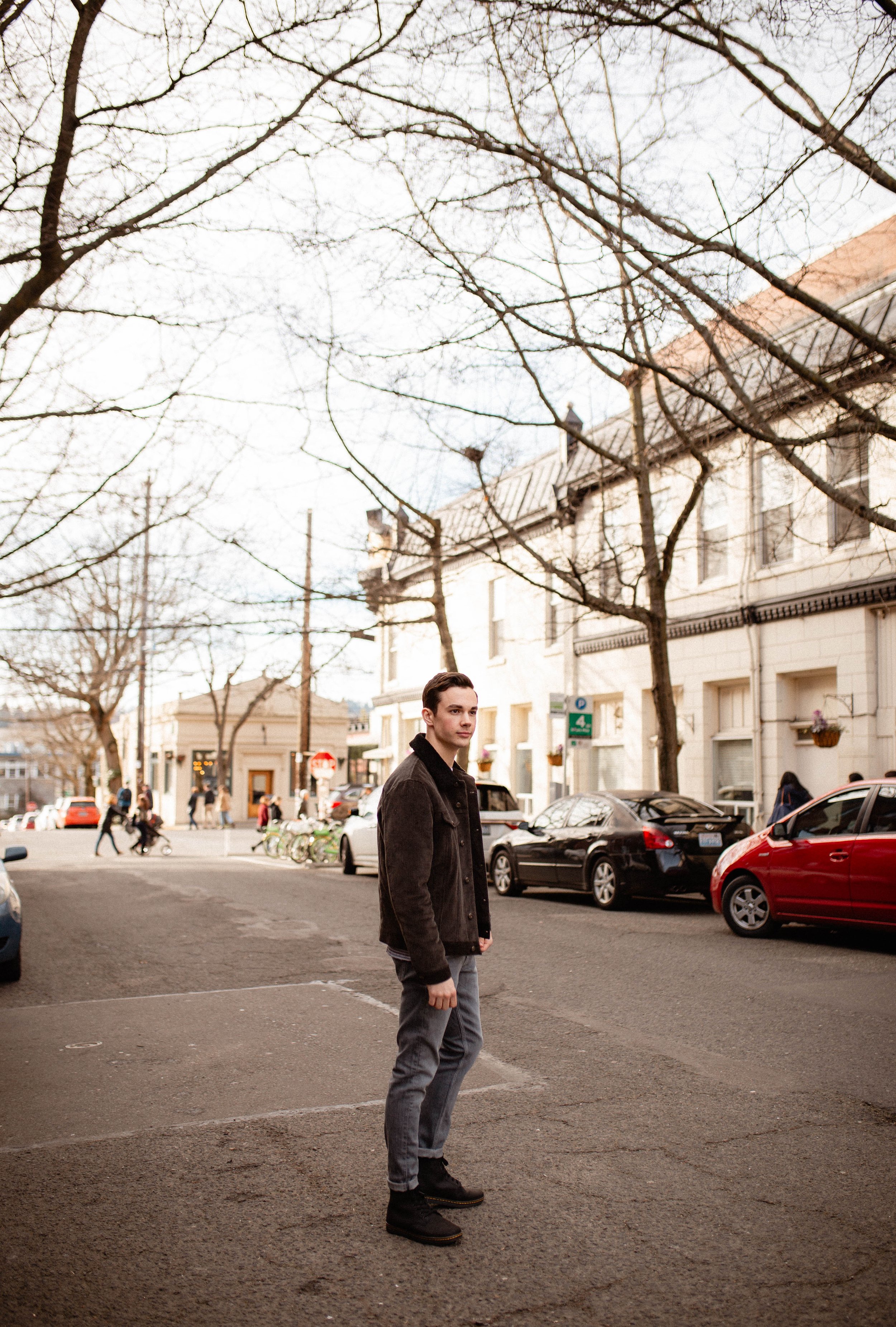 Young man standing on sidewalk in a city street with parked cars and buildings, leafless trees overhead, during daytime. Seattle professional head shot photography