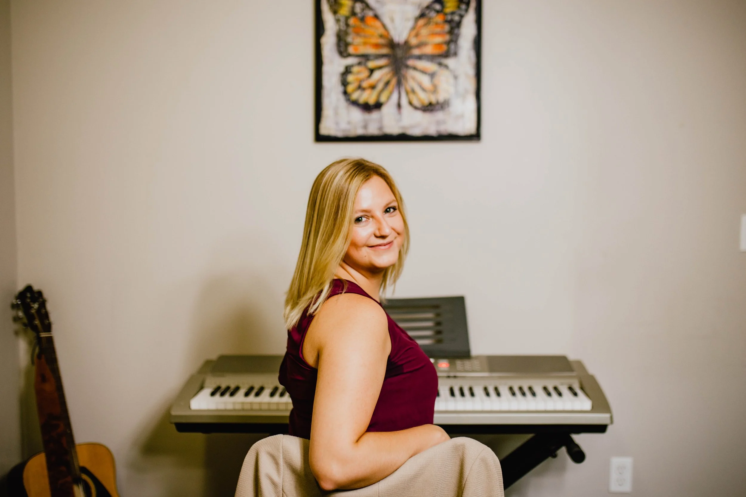 A woman with shoulder-length blonde hair and a sleeveless maroon top sitting in front of a digital keyboard in a room with beige walls and a butterfly painting on the wall. Seattle professional head shot photography