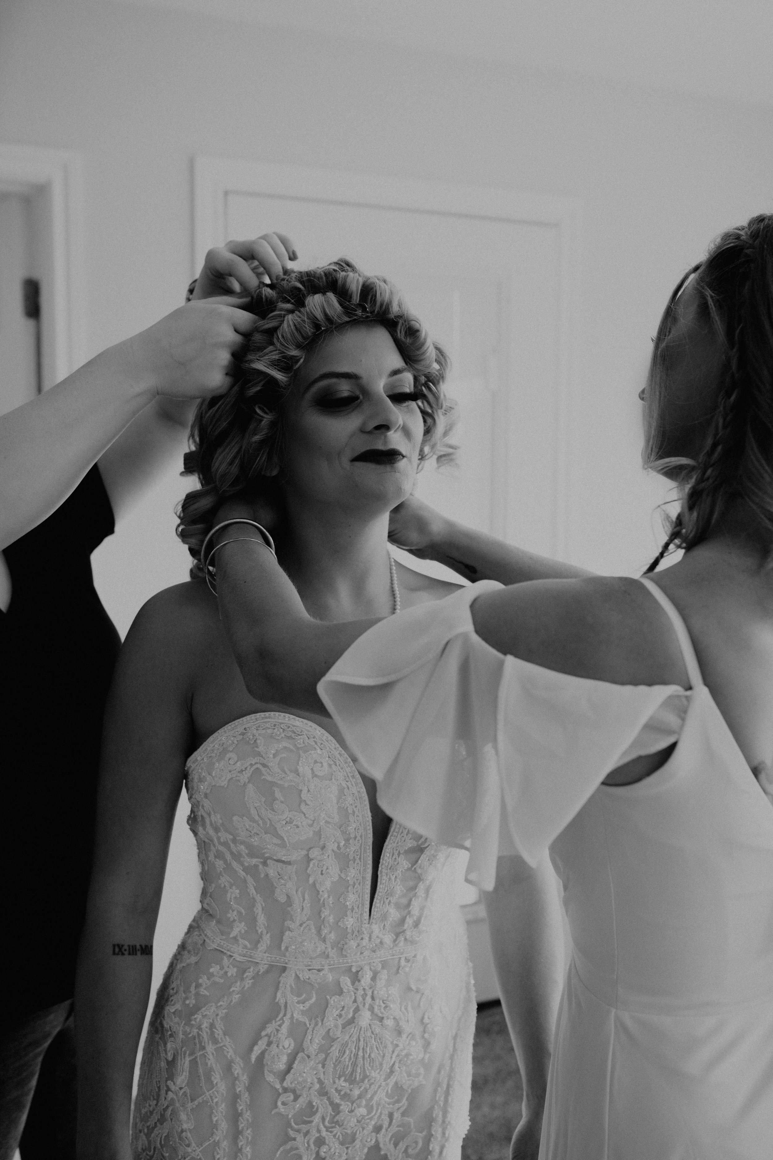 A bride with curly hair and dark makeup gets ready, with helpers adjusting her hair and dress in a room. Long Beach, WA wedding photography.