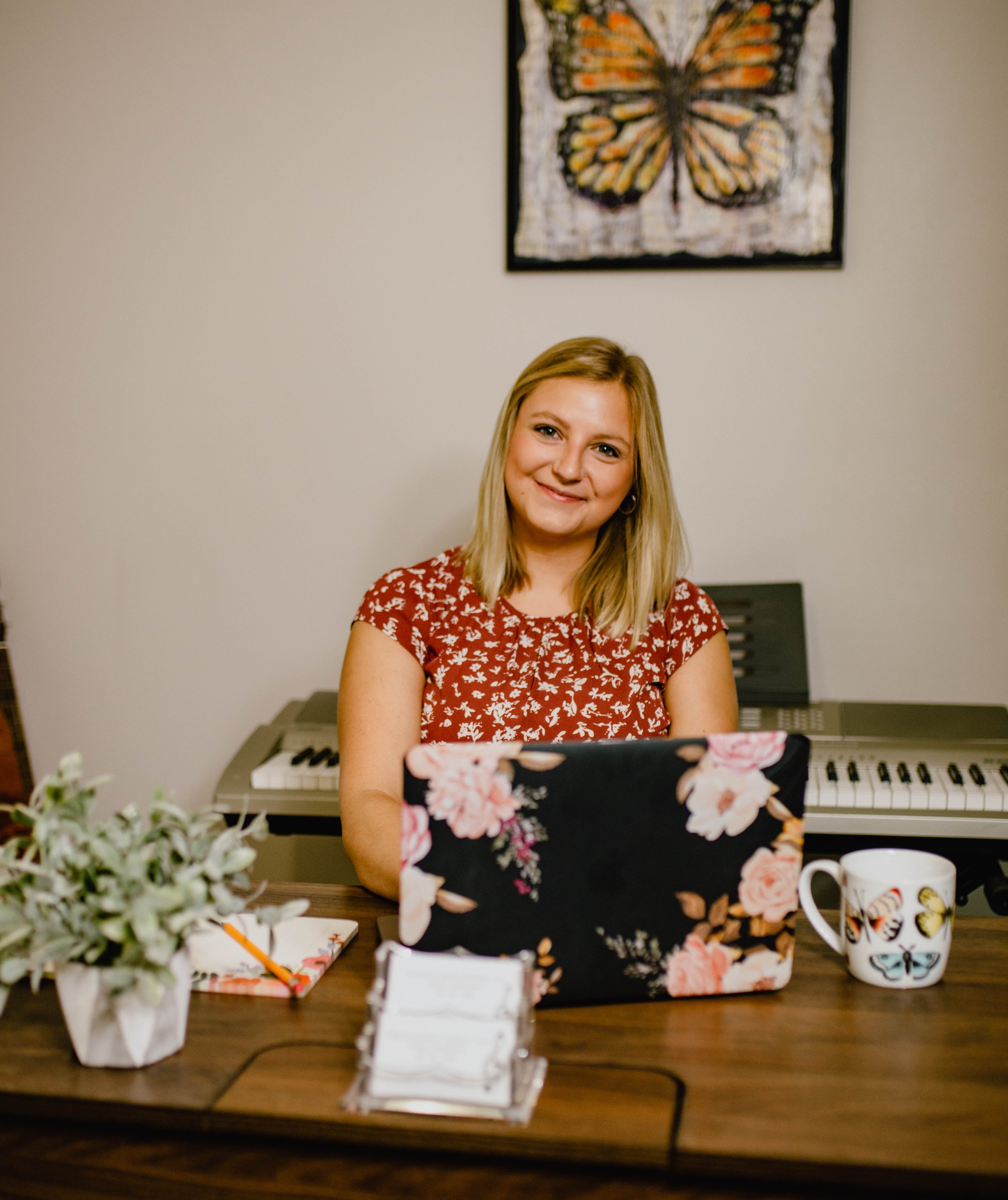 A woman sitting at a desk with a floral laptop, a cup with butterflies, a small plant, and a notebook with an orange pencil, smiling in a room with butterfly artwork on the wall and keyboard in the background. Seattle professional head shot photograp