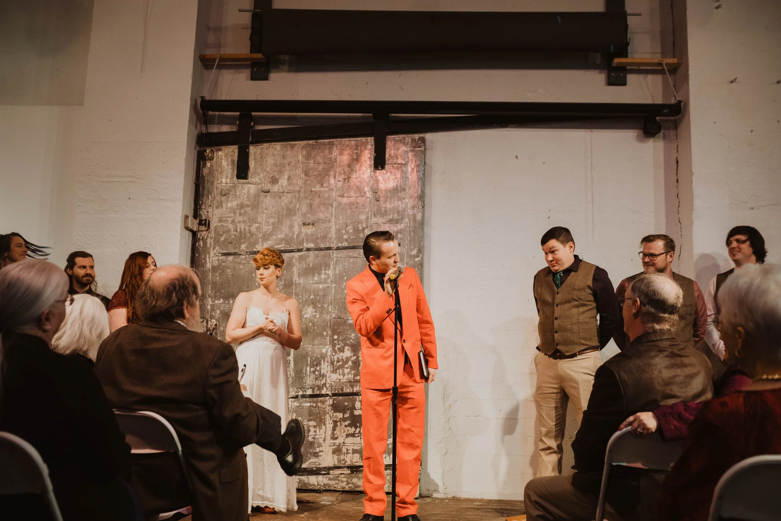 A group of people on stage during a theatrical performance or event. One person in a bright orange suit is speaking into a microphone. The audience seated in front includes older adults and a woman in a white dress stands behind. A rustic metal door 