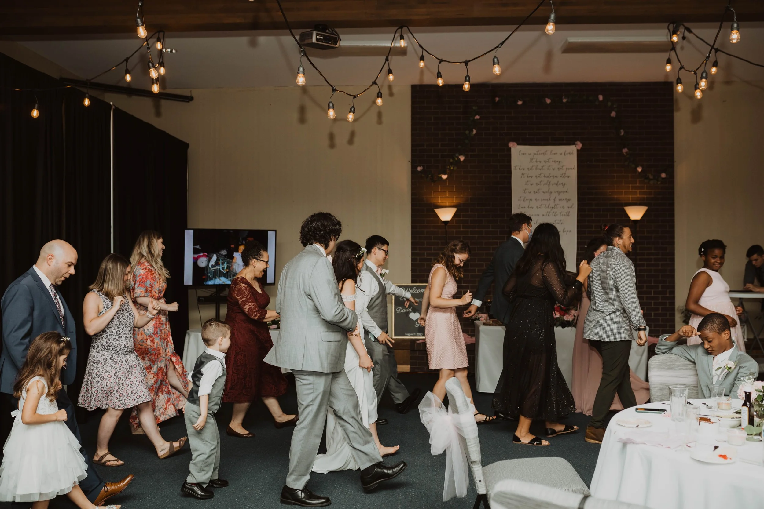 Guests at a wedding reception dancing in a line indoors, with string lights hanging from the ceiling and a brick wall with floral decorations in the background Seattle, WA wedding photography. .