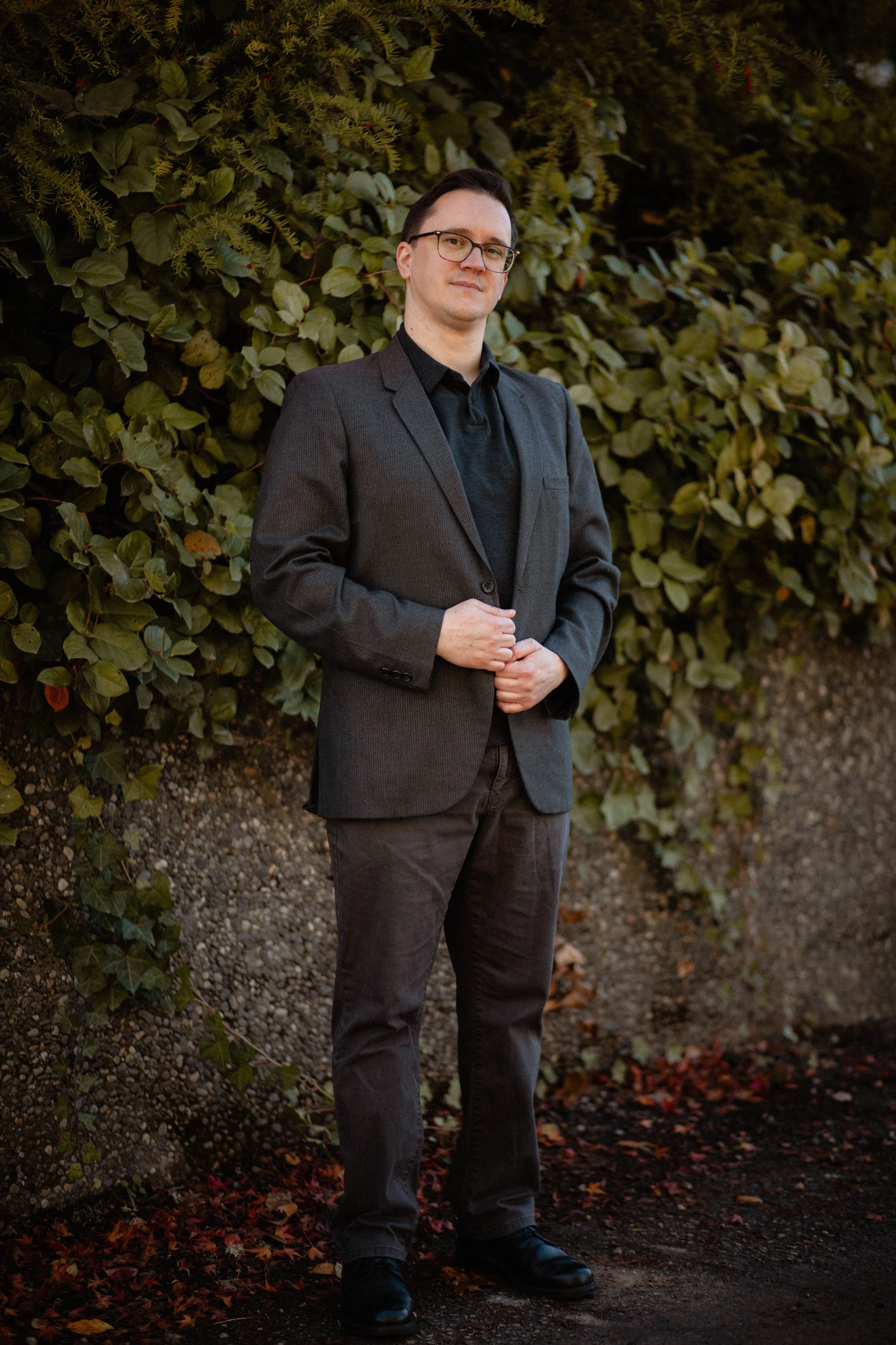 A man in a dark suit standing outdoors near a hedge with dark soil and fallen leaves at his feet. Seattle professional head shot photography