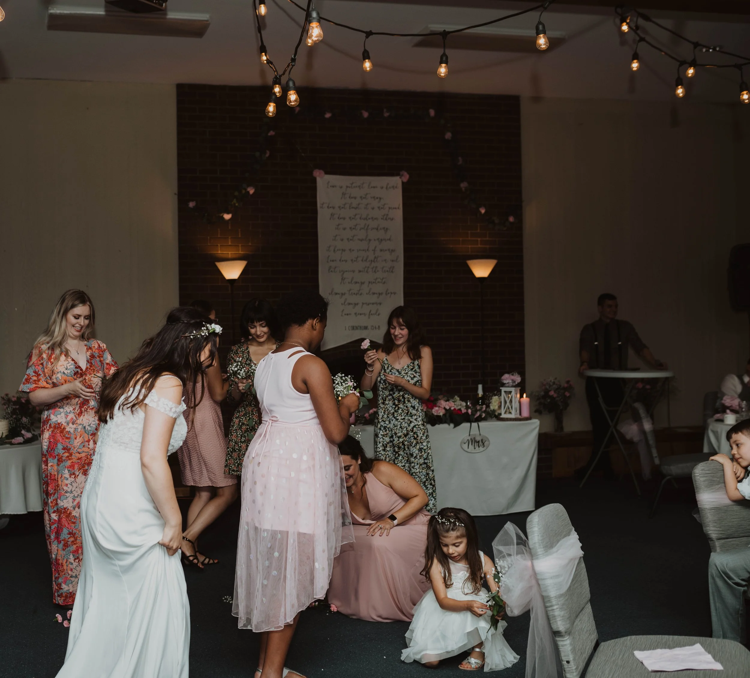 Group of women and children at a wedding reception, with some women in dresses and a flower girl holding flowers, standing near tables decorated with flowers and candles, in a room with hanging string lights and a brick wall. Seattle, WA wedding phot