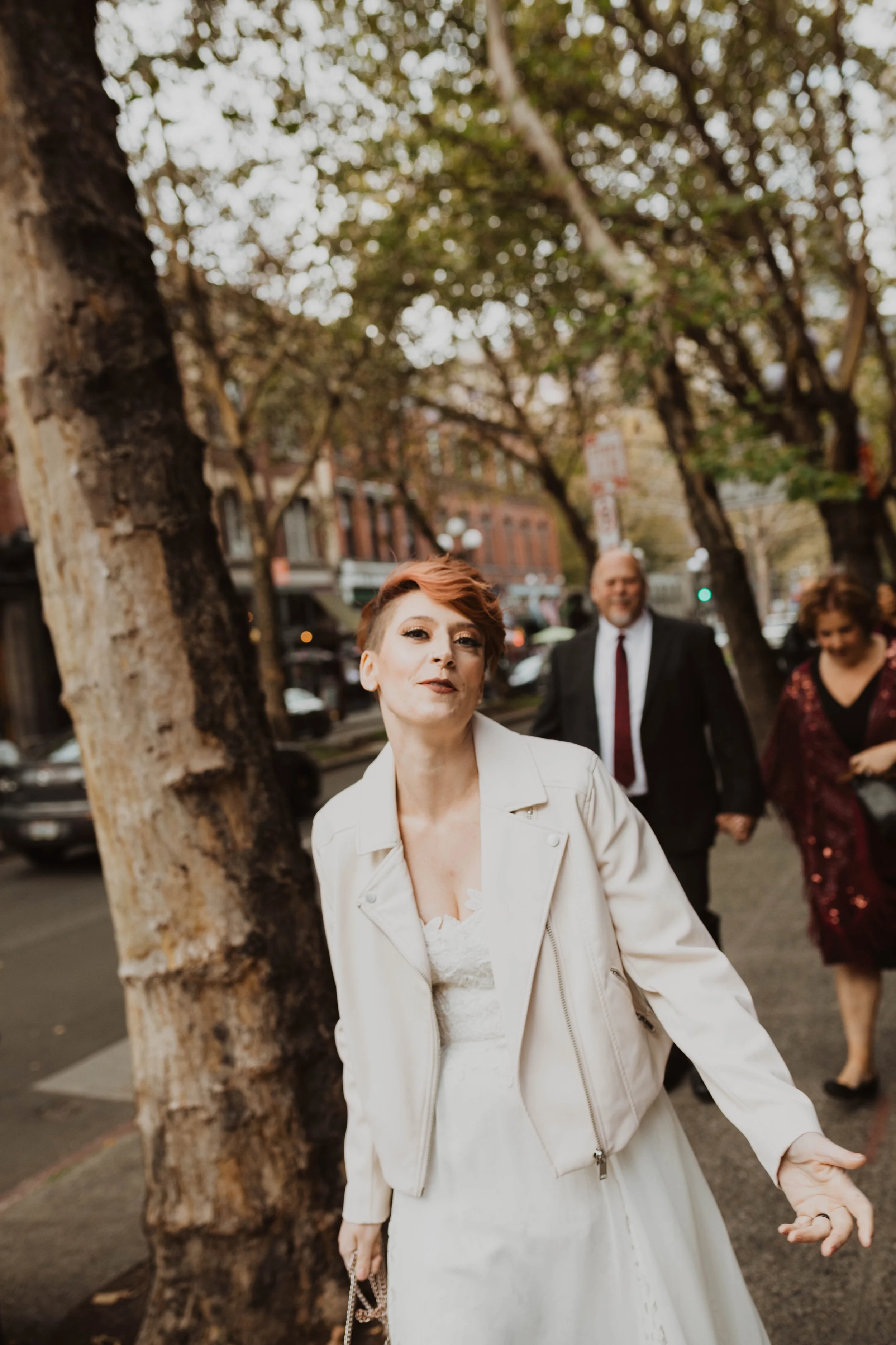 A woman with short red hair in a white dress and white jacket walking on a city sidewalk, with two men and another woman in the background, trees lining the street. Pioneer Square, Seattle, WA wedding photography.