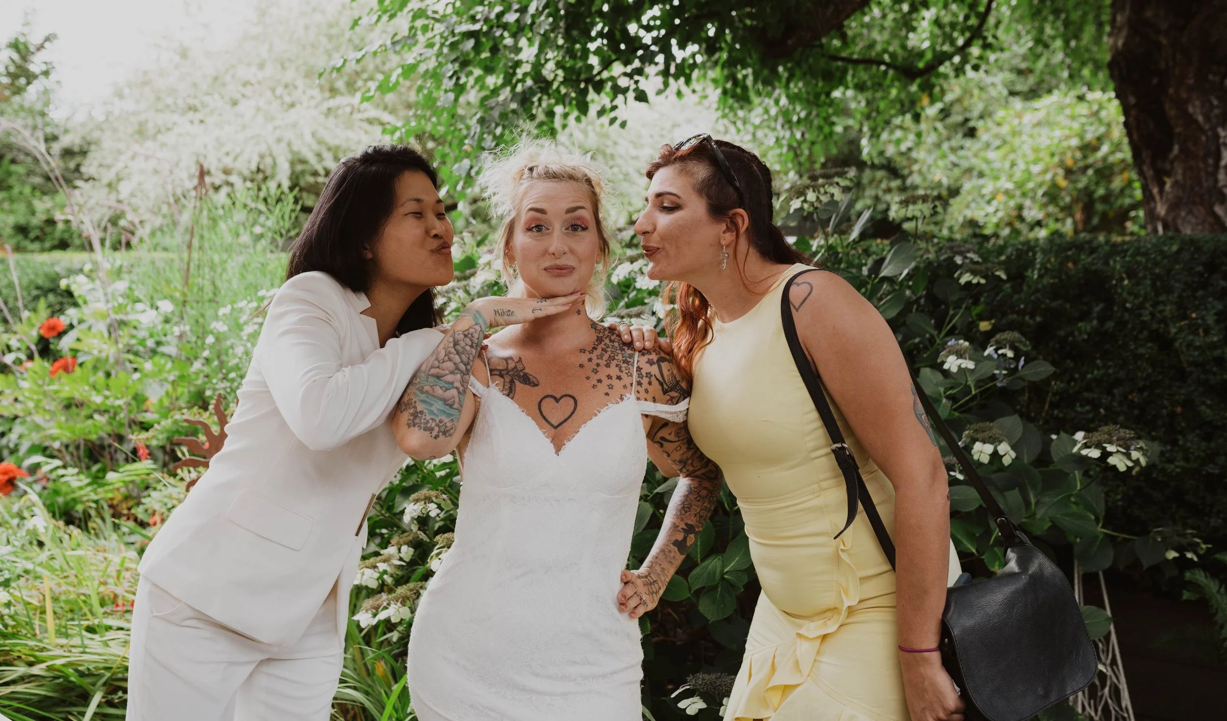 Three women, one in a white dress, and two in casual yellow and white outfits, are posing playfully and affectionately outdoors among greenery and flowers. Seattle, WA wedding photography.