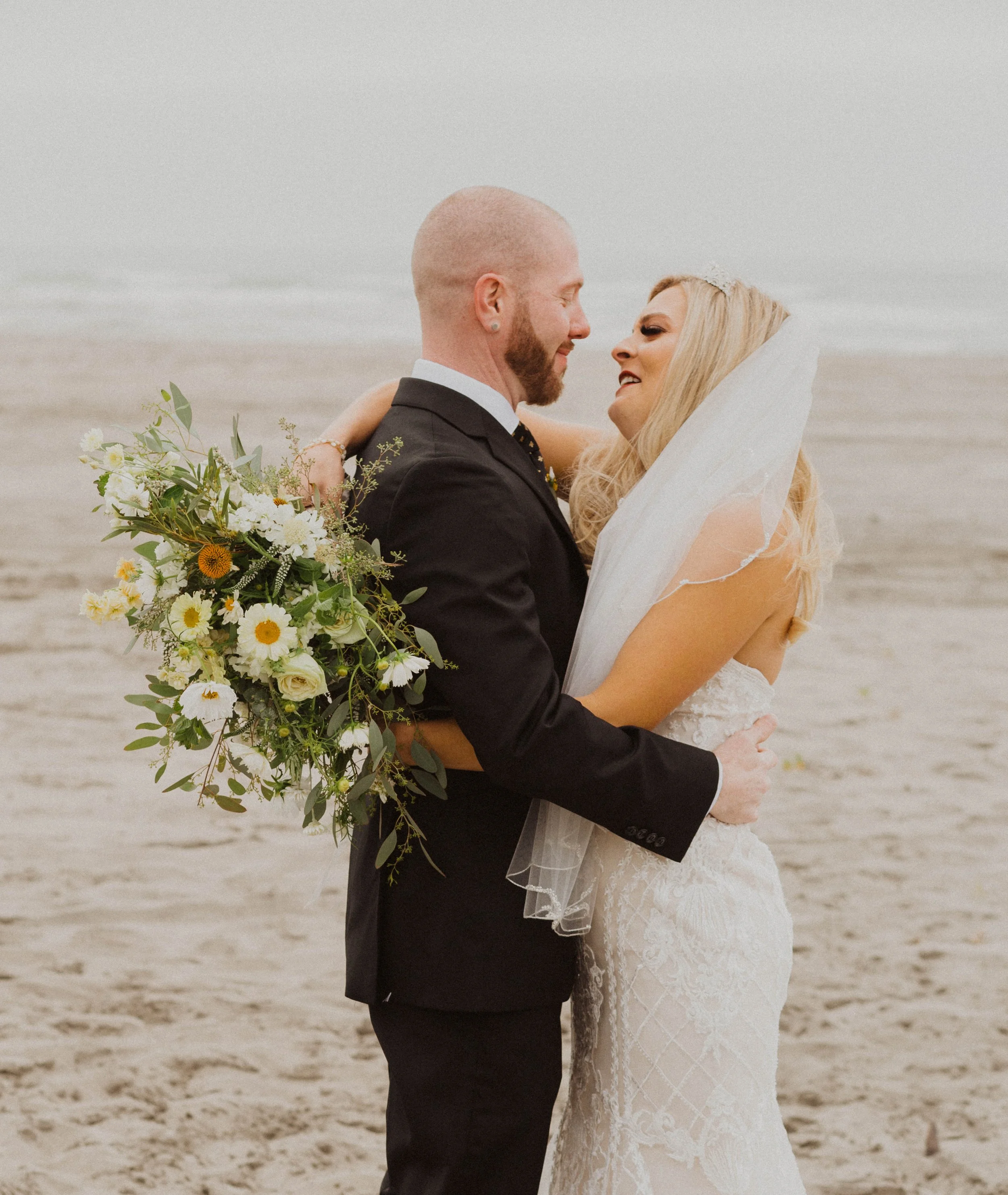 A bride and groom are standing close together on a beach, gazing into each other's eyes. The bride is holding a large bouquet of flowers, and the groom is wearing a black suit. The bride has a long veil and a wedding dress with lace details.