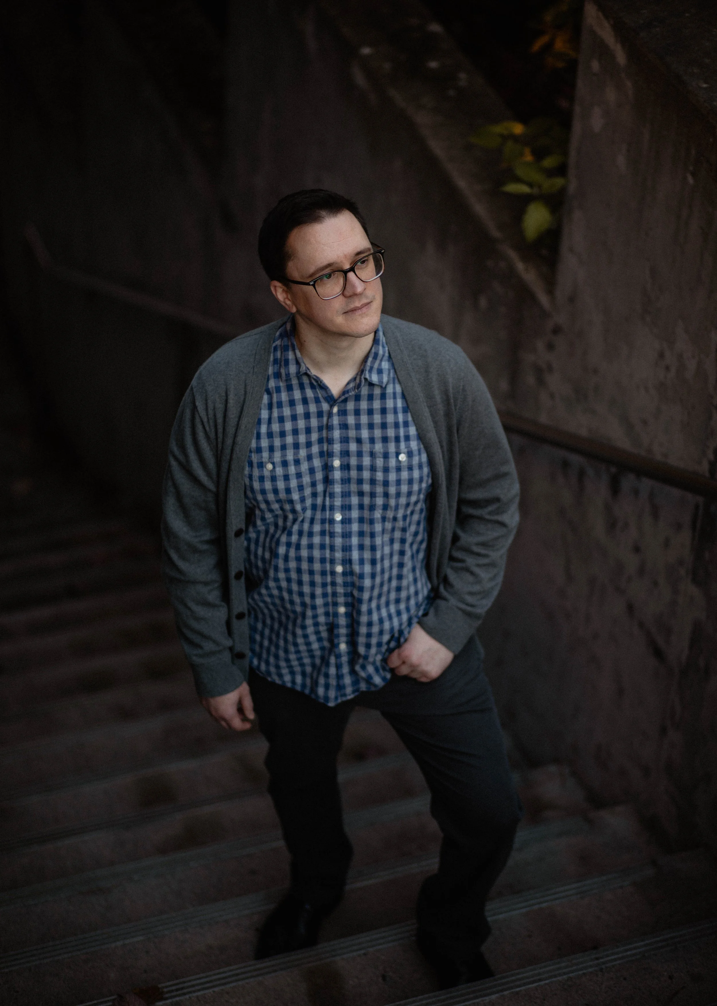 A man with glasses, dark hair, and light skin wearing a checkered blue shirt, gray cardigan, and black pants standing on a dimly lit staircase with stone walls and a handrail. Seattle professional head shot photography