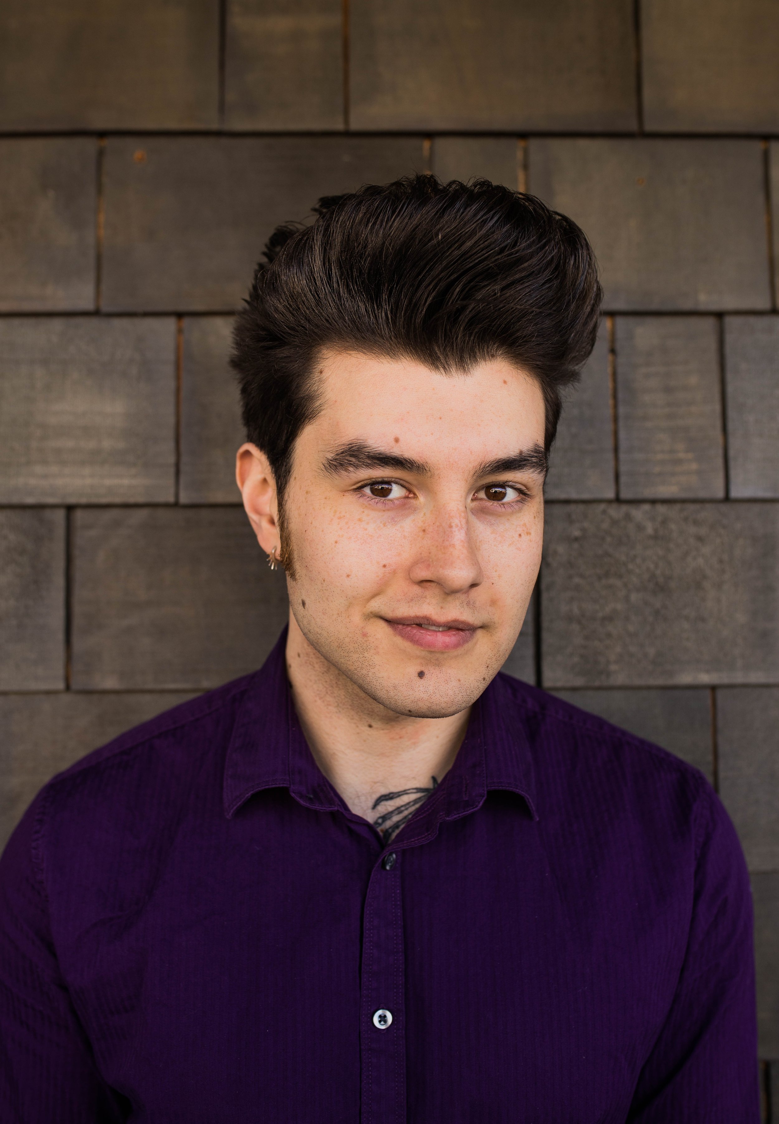 A young man with dark hair styled upward, wearing a dark purple shirt, standing against a wooden wall, looking at the camera with a slight smile. Seattle professional head shot photography