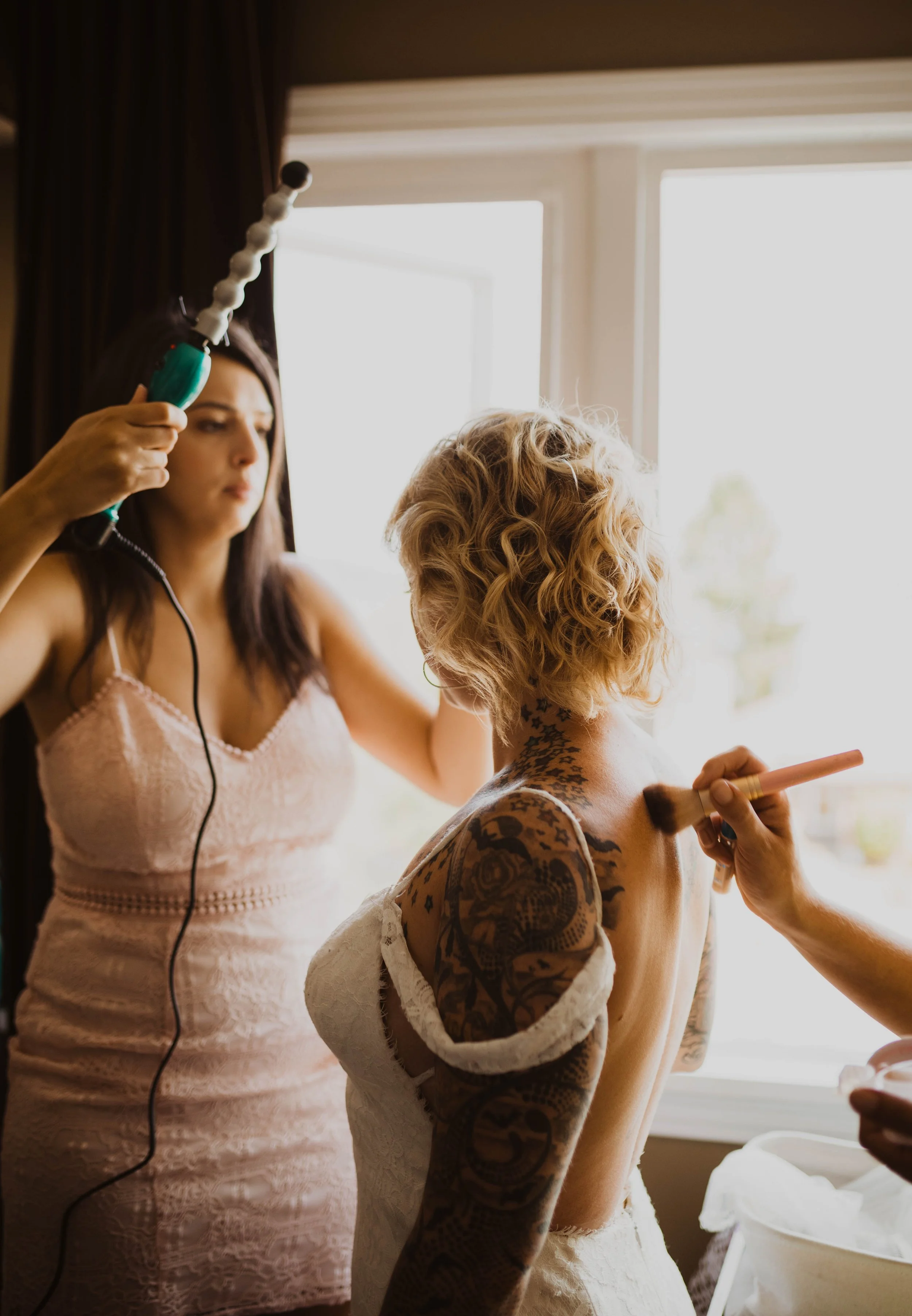 A woman in a white dress is having her makeup done while another woman with dark hair uses a heated tool on her hair in front of a window with natural light. Seattle, WA wedding photography.
