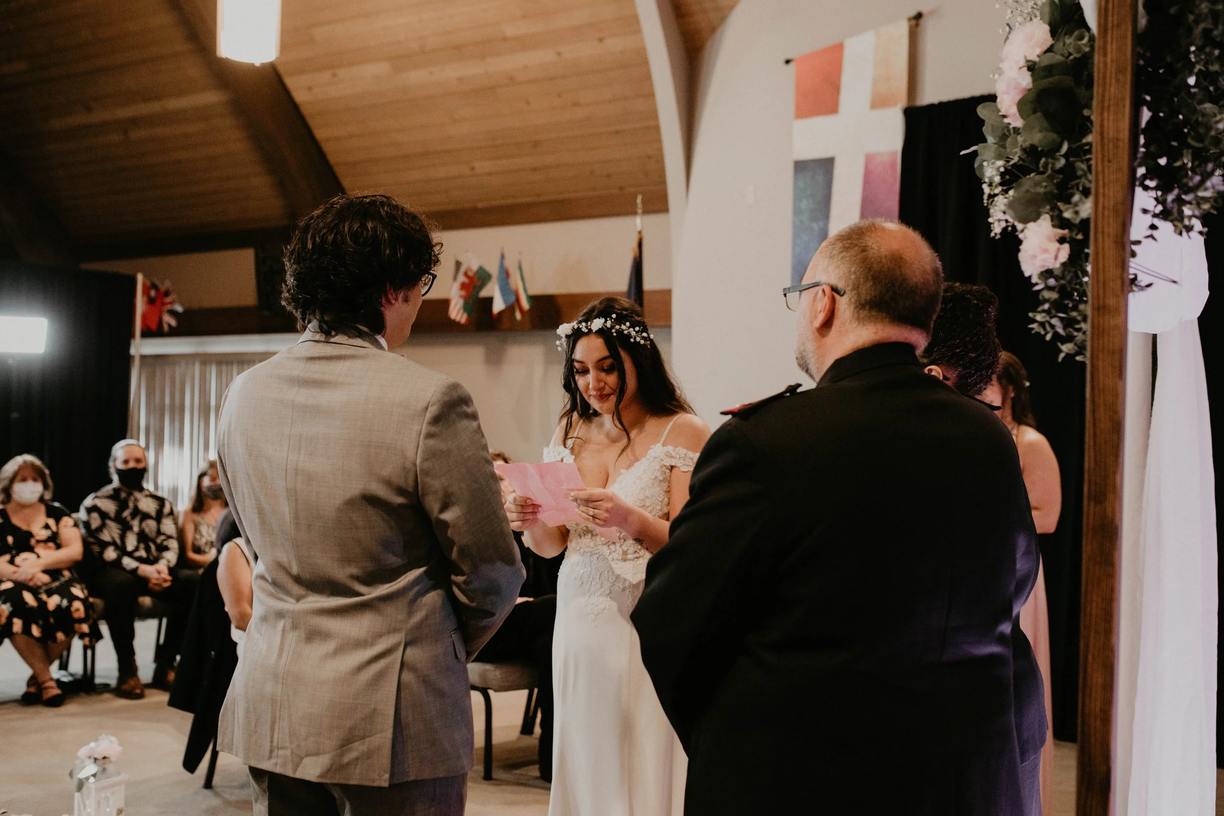 A bride reading vows during her wedding ceremony, standing under a floral arch with a groom and officiant in a decorated church or event hall, with guests seated in the background. Seattle, WA wedding photography.