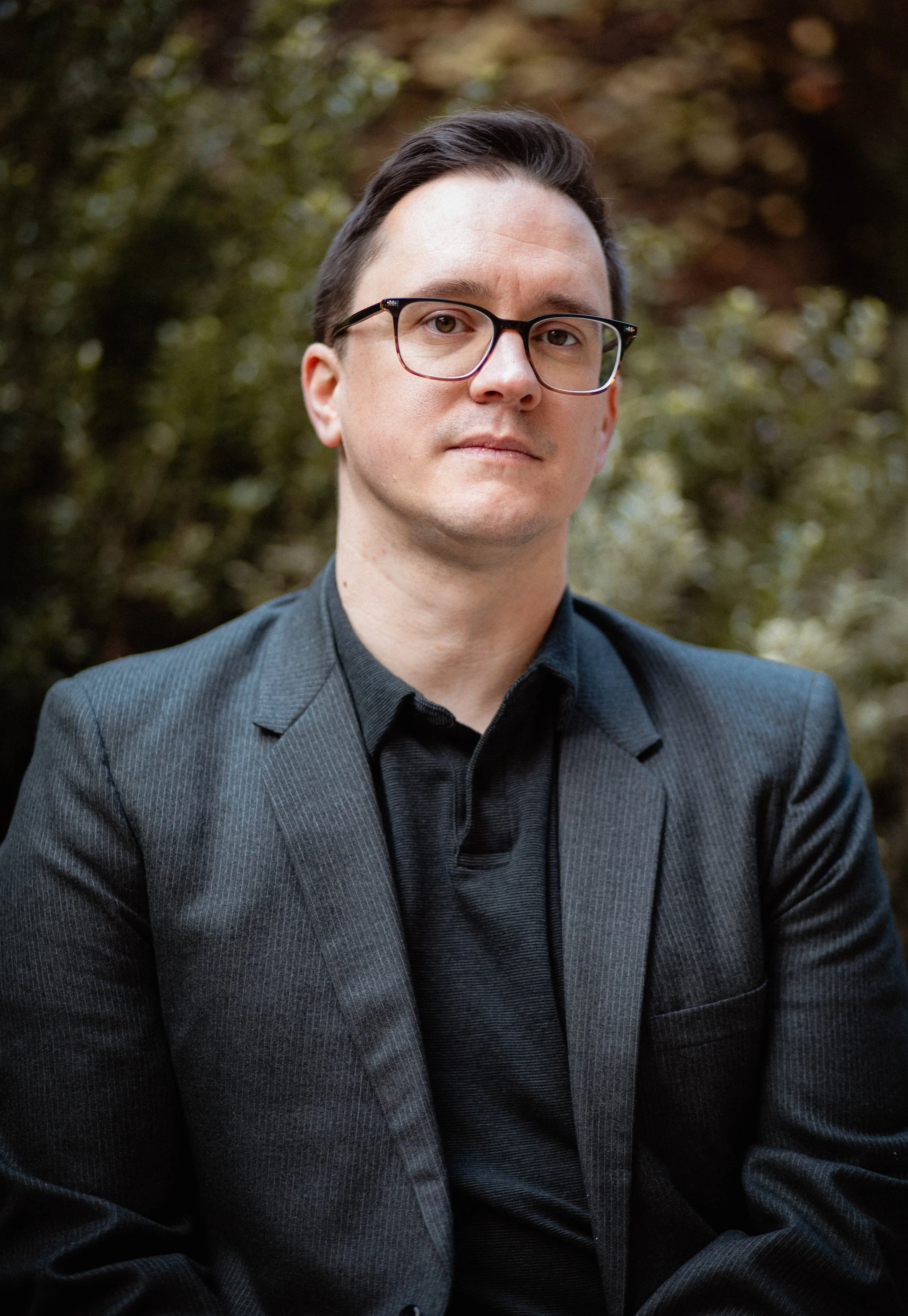 A man wearing glasses in a black suit and black shirt standing outdoors with blurred trees in the background. Seattle professional head shot photography