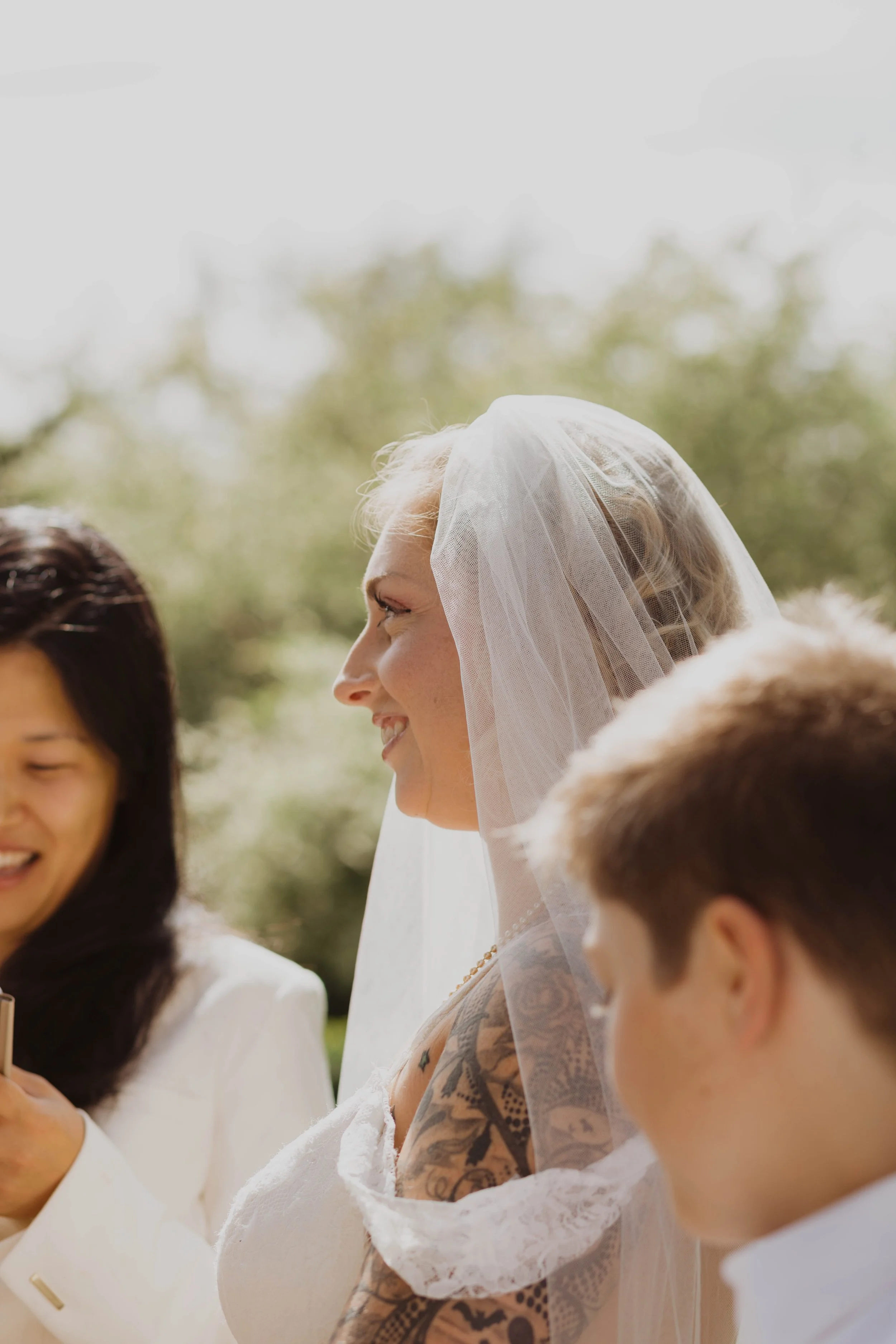A bride in a wedding dress and veil smiling, with two people nearby, outdoors with blurred green trees background. Seattle, WA wedding photography.