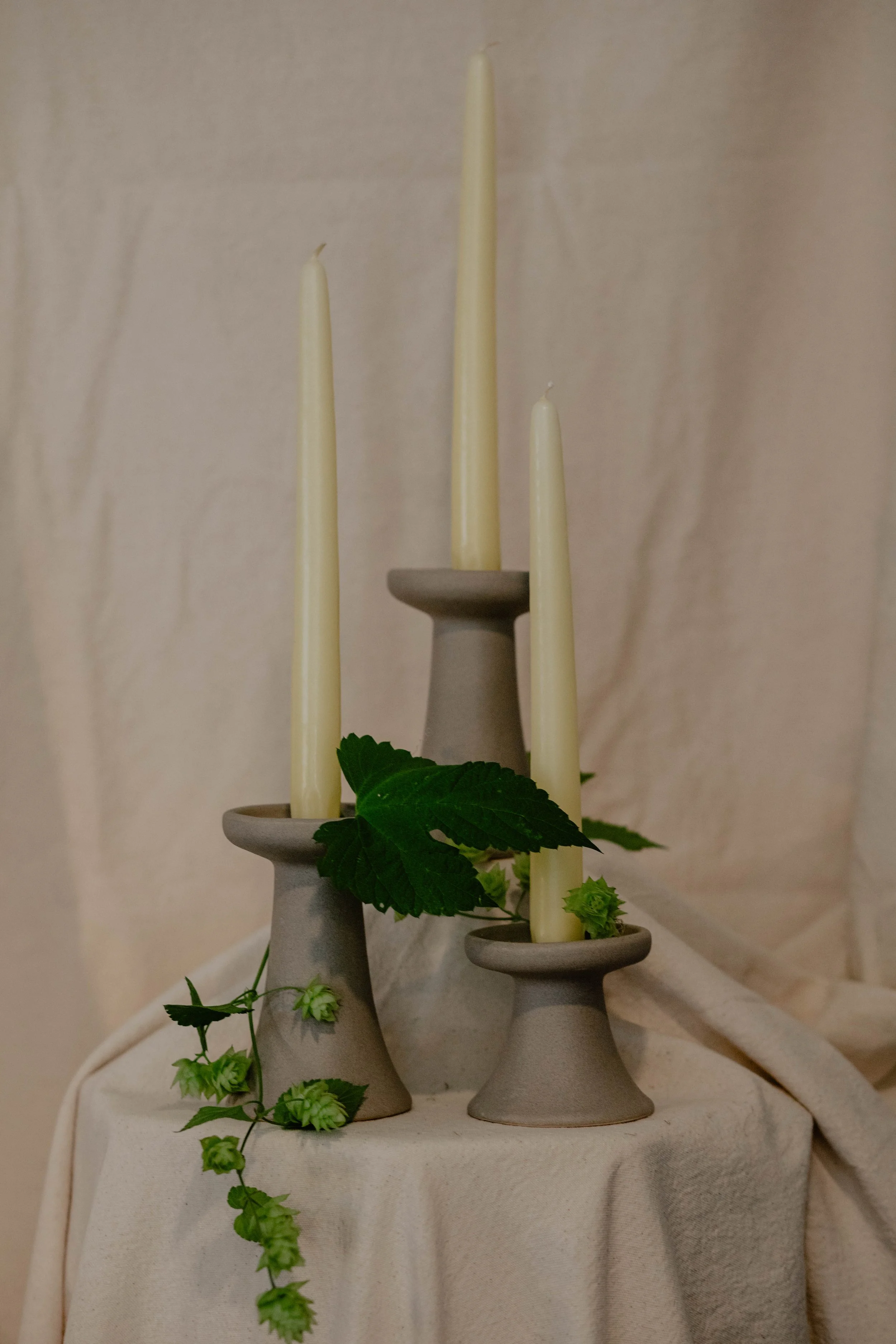 Three gray candle holders of varying heights with cream-colored candles, decorated with green leaves and hop plant, placed on a cream-colored cloth-covered surface against a light background.  Seattle product photography