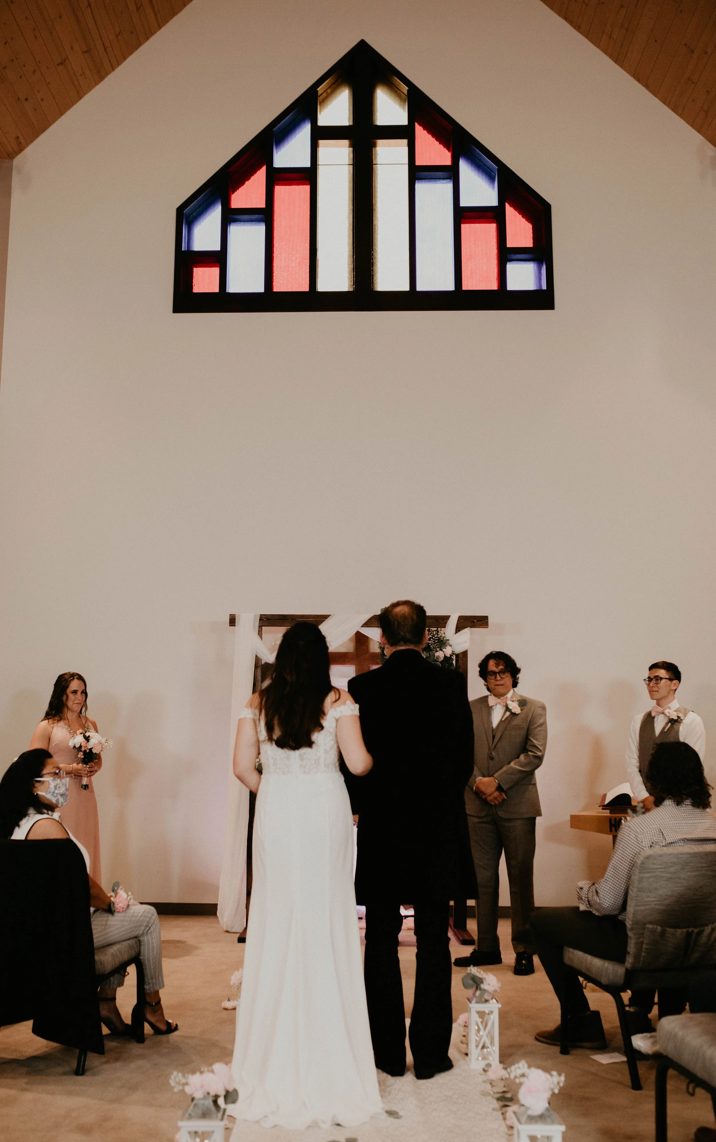 A couple getting married during a wedding ceremony inside a church, with guests seated on either side, a officiant, and floral decorations. A colorful stained glass window with red, blue, white, and black sections is above them. Seattle, WA wedding p