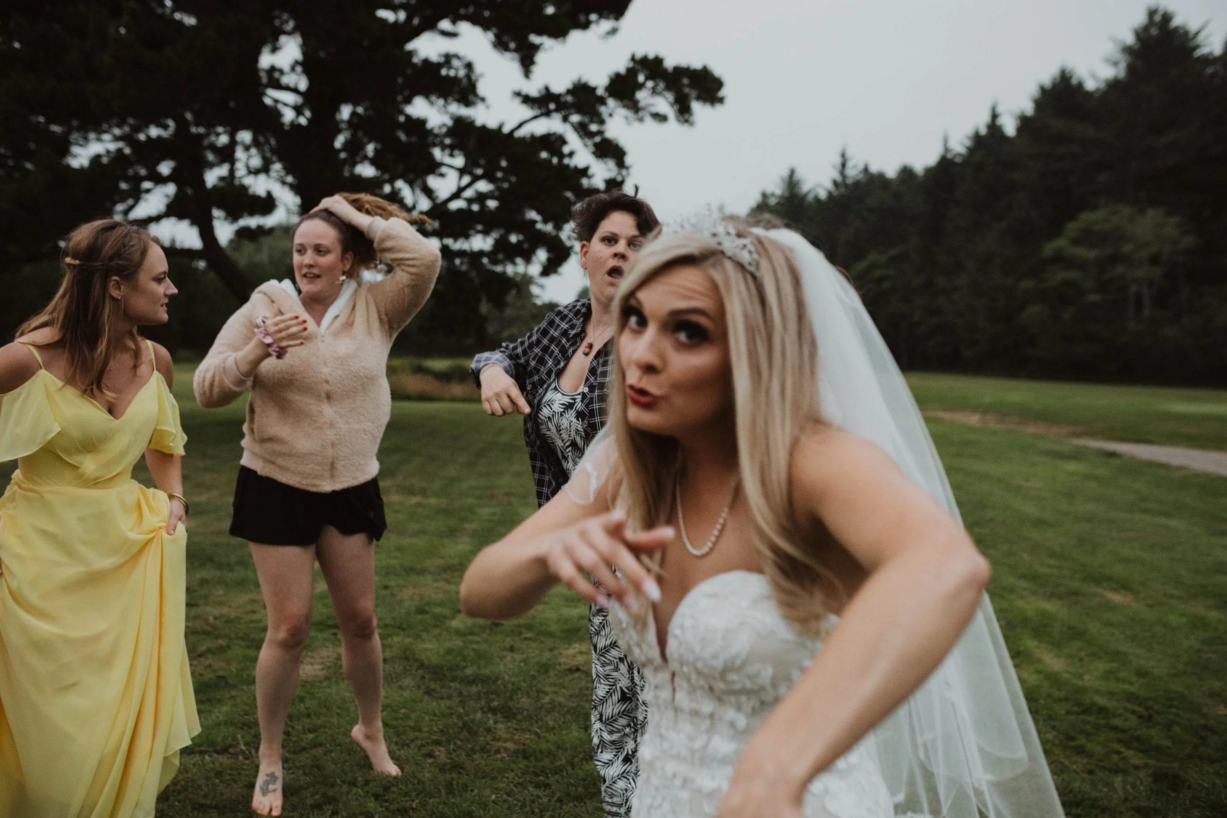 A bride in a wedding dress and veil making a playful gesture, standing outdoors with three women behind her talking and laughing, on a grassy field with trees in the background. Long Beach, WA wedding photography.