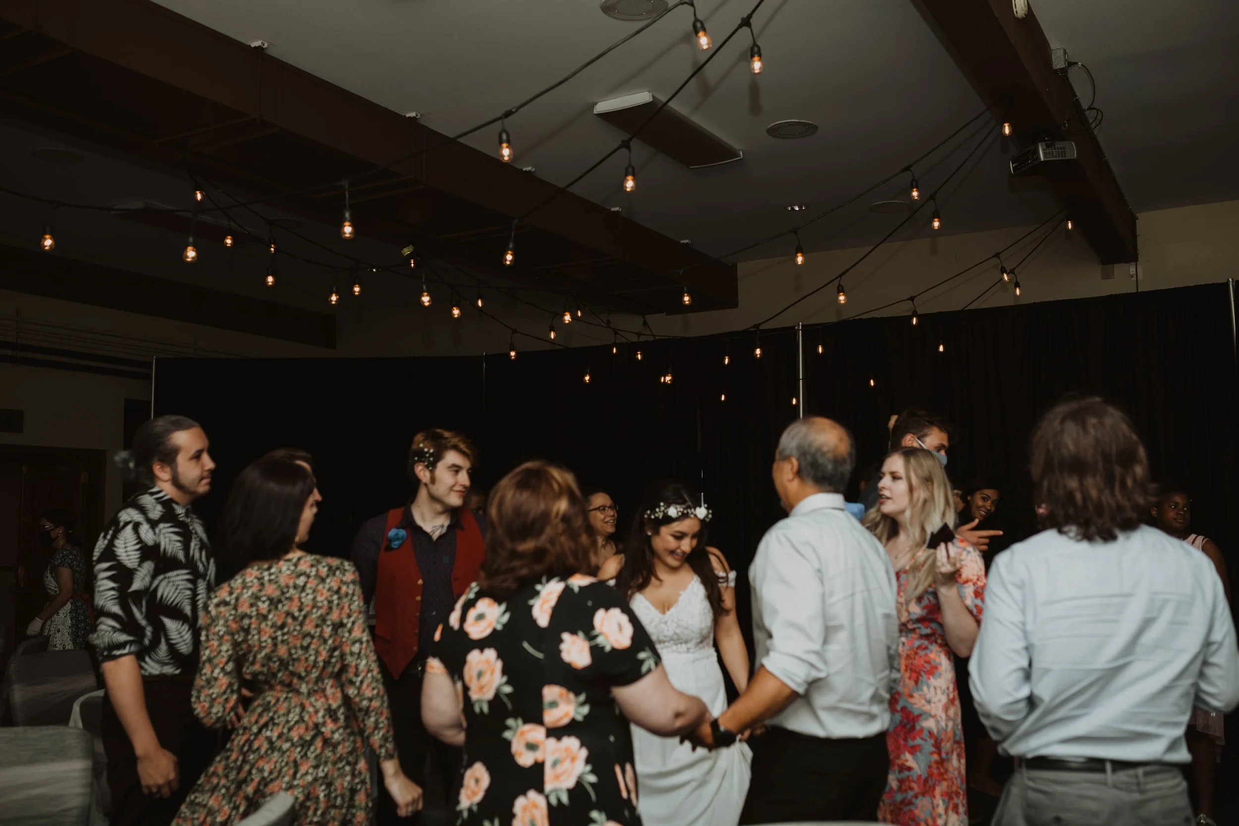People dancing in a circle at a celebration or wedding in a dimly lit room with string lights overhead. Seattle, WA wedding photography.