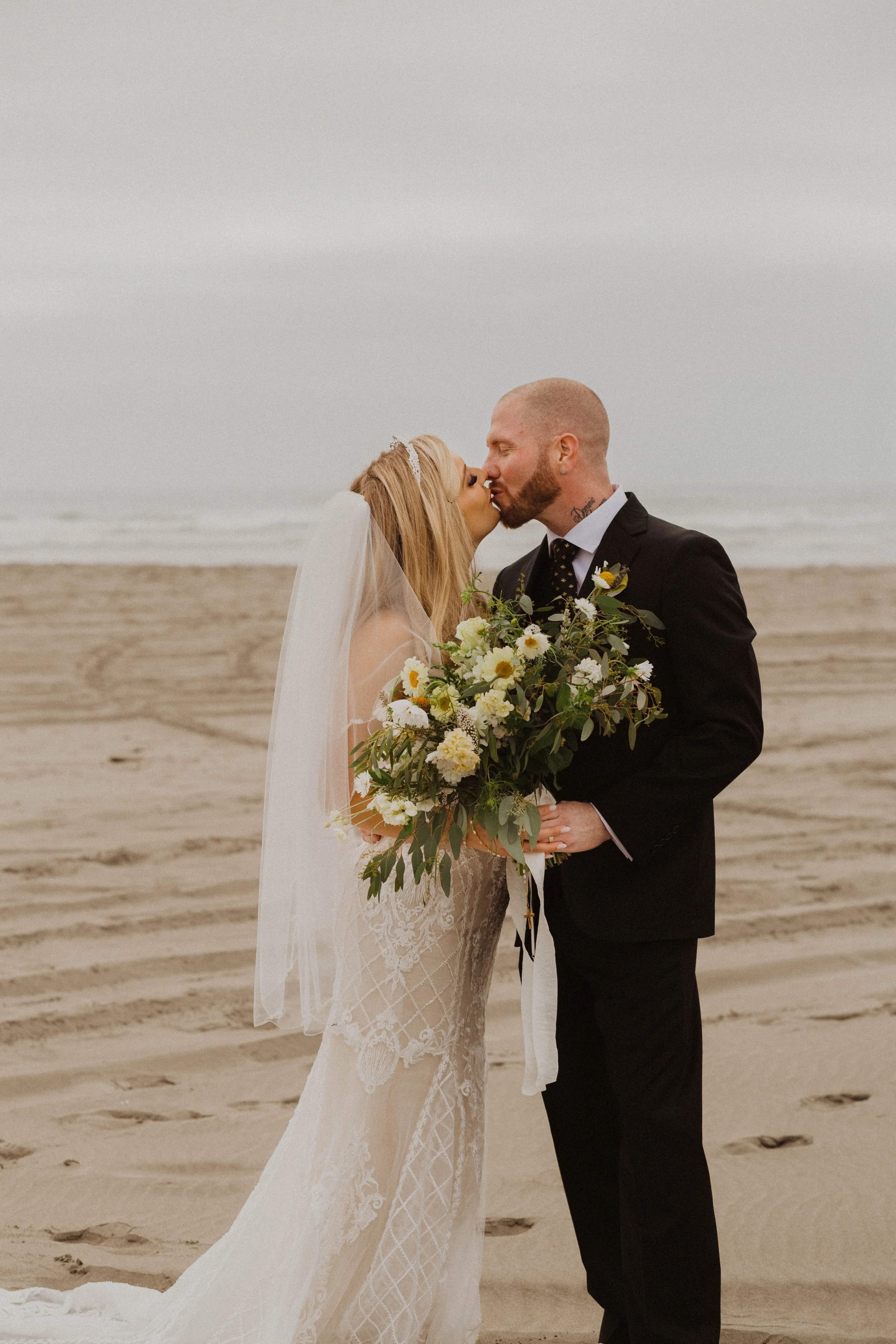 Bride and groom kissing on the beach with the bride holding a large bouquet of white and yellow flowers. Long Beach, WA wedding photography.