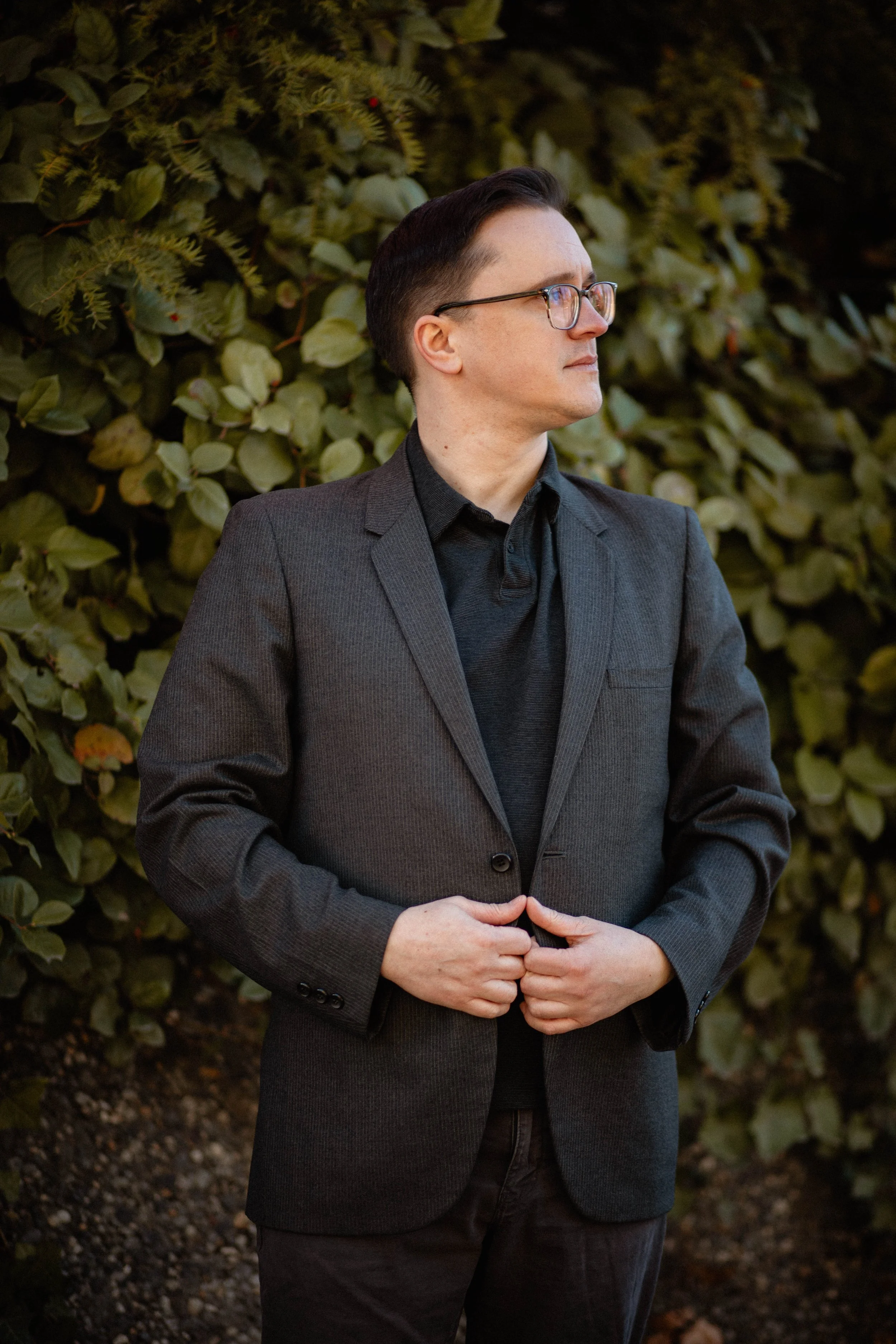A man in a dark gray blazer with glasses looking to the right, standing outdoors in front of green foliage. Seattle professional head shot photography