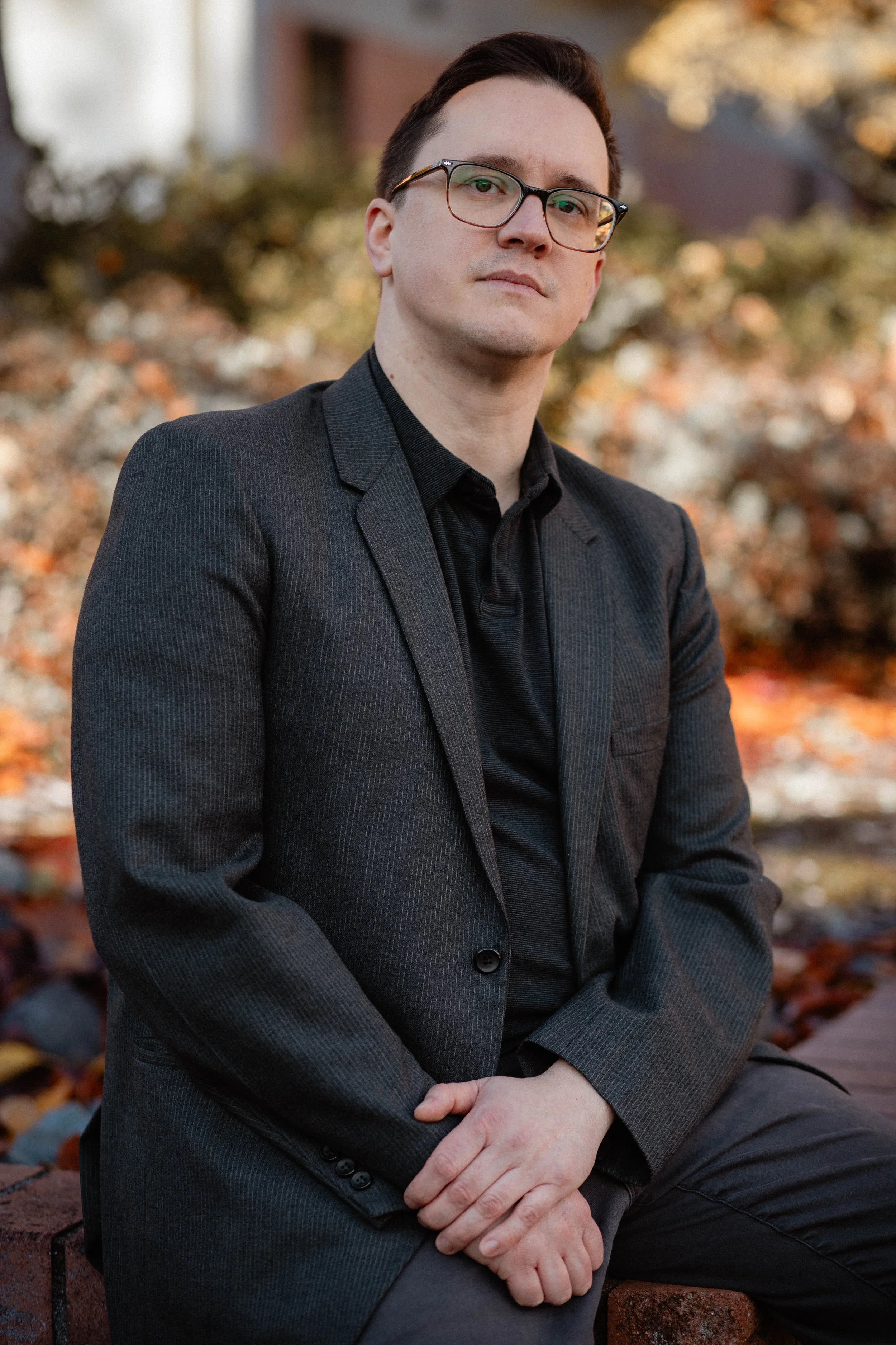 A young man with dark hair and glasses wearing a black blazer sitting outdoors during autumn. Seattle professional head shot photography