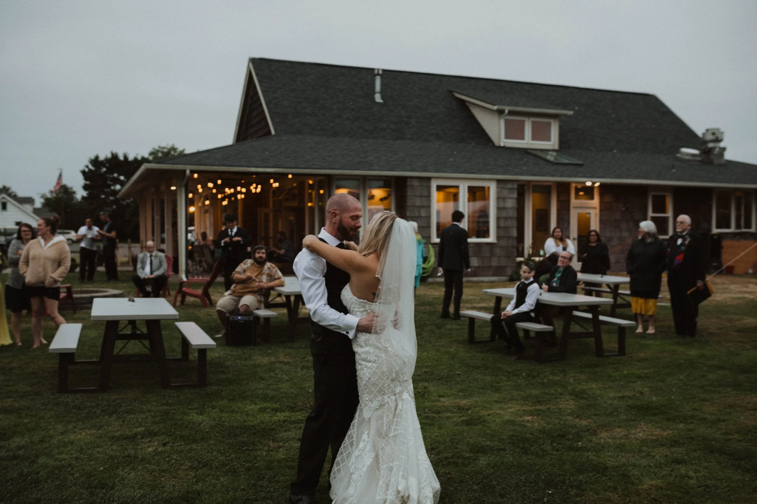 A bride and groom dancing outdoor at their wedding, surrounded by guests sitting and standing at picnic tables and a house decorated with string lights in the background during evening. Long Beach, WA wedding photography.