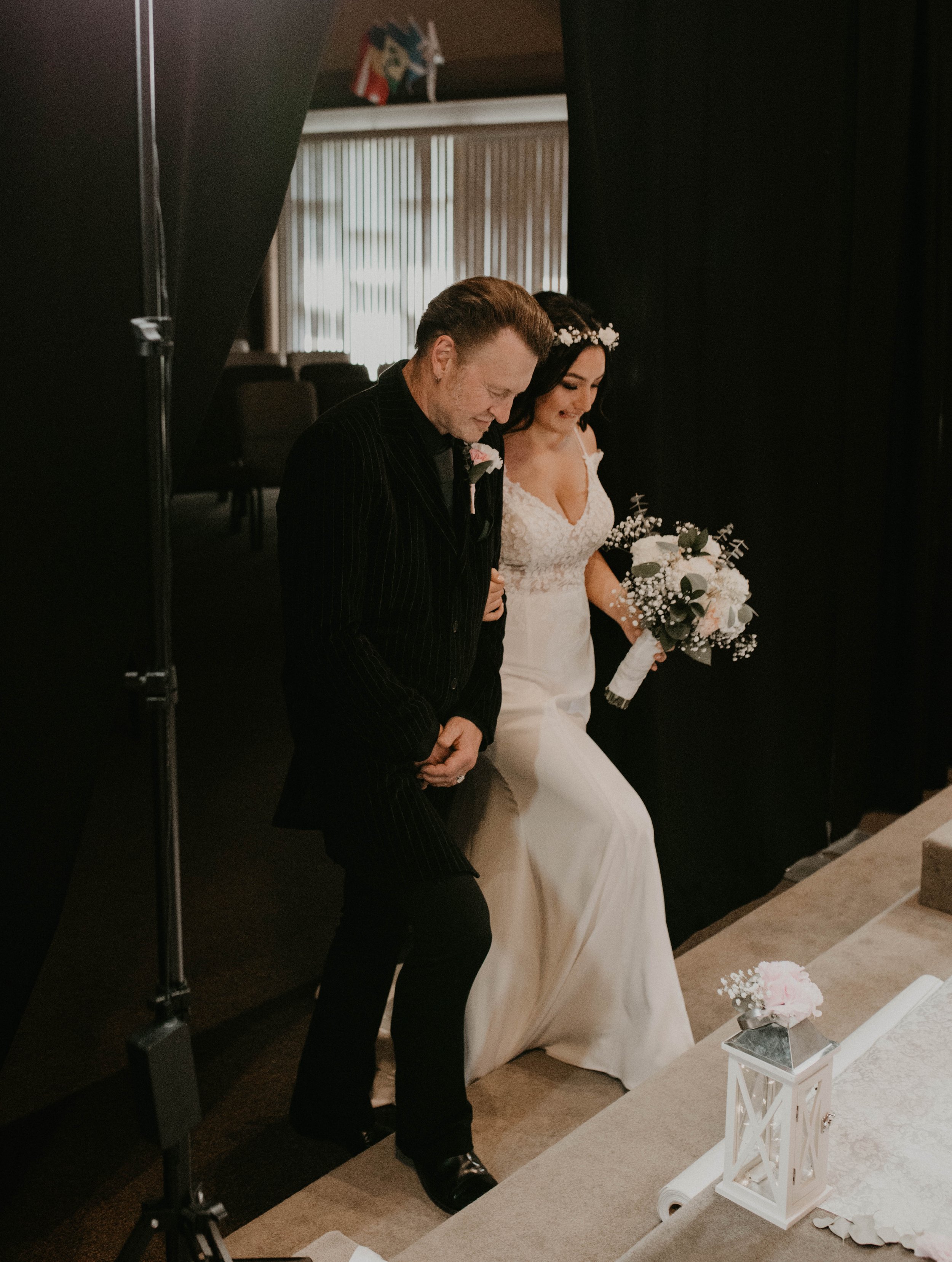 A bride in a white wedding dress walking down the aisle with an older man, likely her father, during a wedding ceremony. The bride holds a bouquet of flowers, and the scene is set indoors with black curtains and a small table with a lantern and flowe