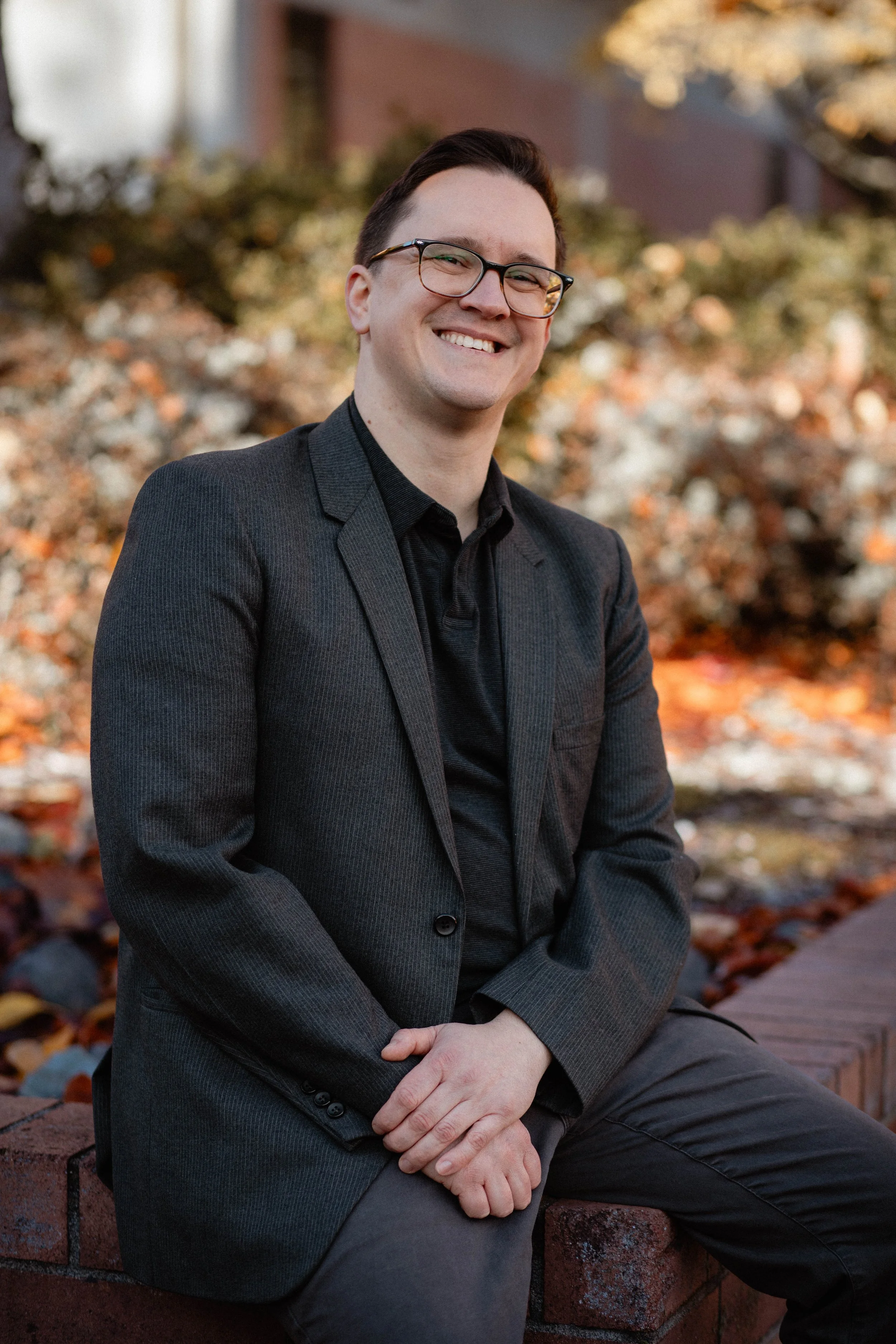 A man in a black blazer and glasses sitting on a brick wall outdoors, smiling with autumn leaves in the background. Seattle professional head shot photography