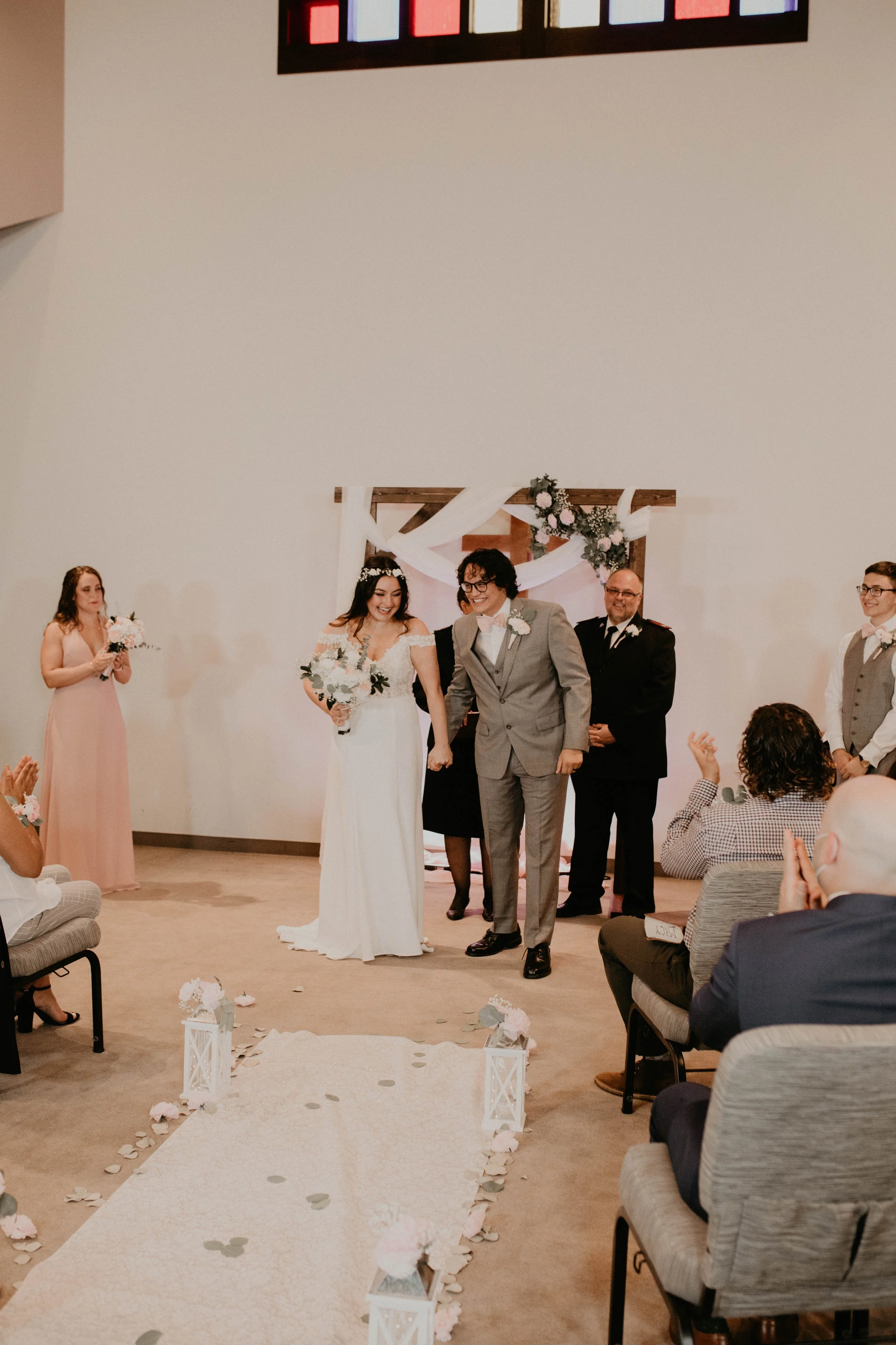 A wedding ceremony taking place indoors with a bride and groom walking down the aisle, surrounded by bridesmaids and groomsmen. Guests are seated and applauding, and the aisle is decorated with lanterns, flowers, and rose petals.