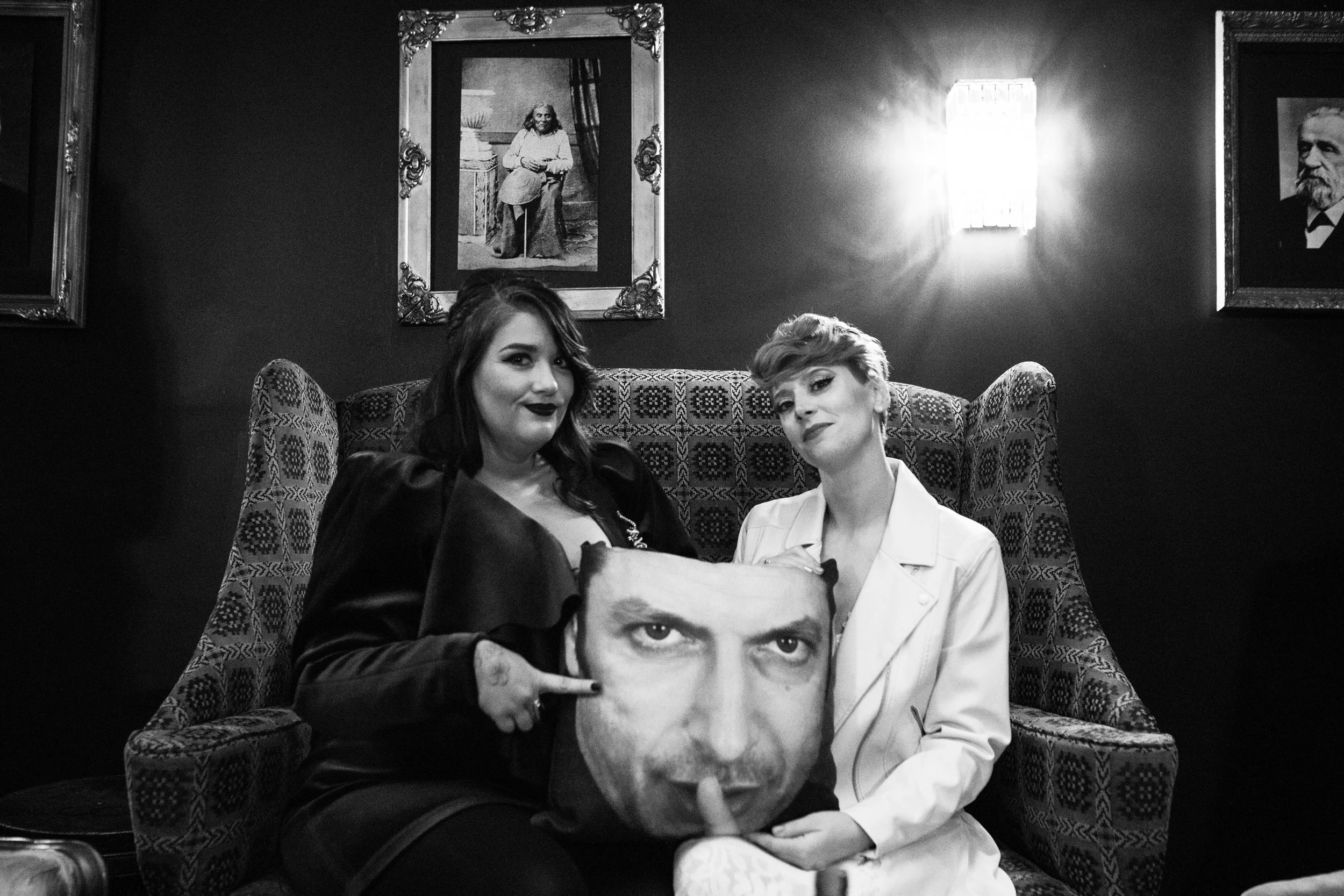Two women sitting on a patterned armchair in a dimly lit room with framed paintings on the wall. One woman holds a pillow with a large, printed face, and the other woman points at the pillow. Pioneer Square, Seattle, WA wedding photography.