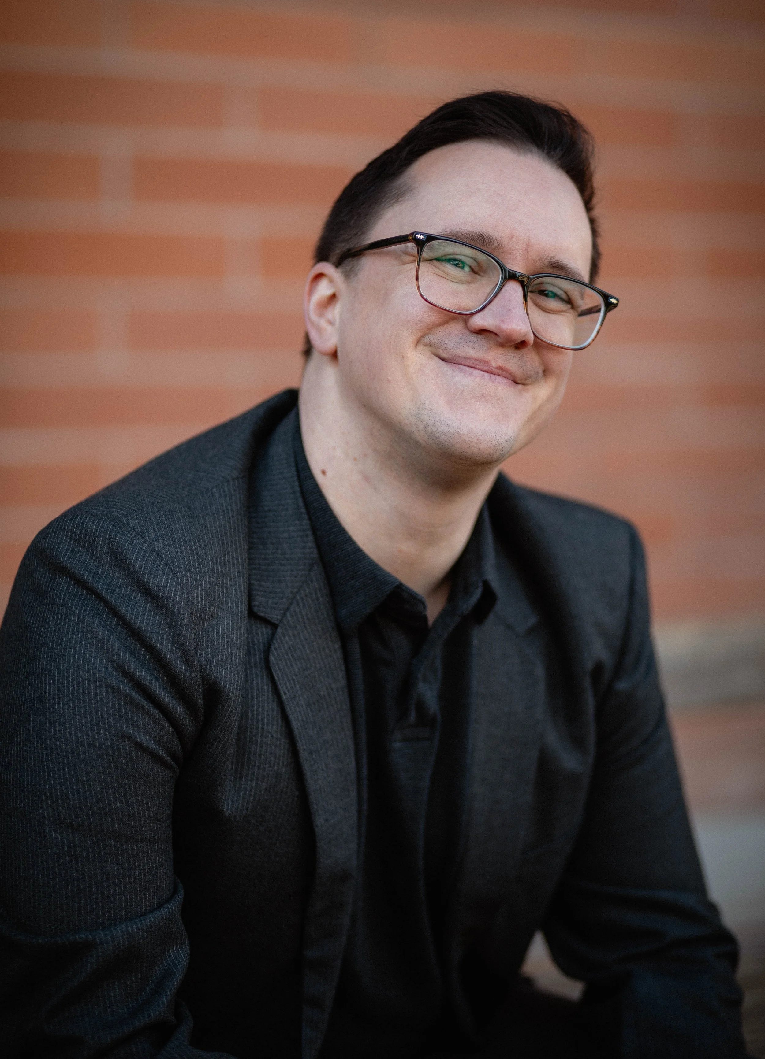 A man with dark hair, glasses, and a slight smile, wearing a black blazer and shirt, sitting in front of a brick wall. Seattle professional head shot photography