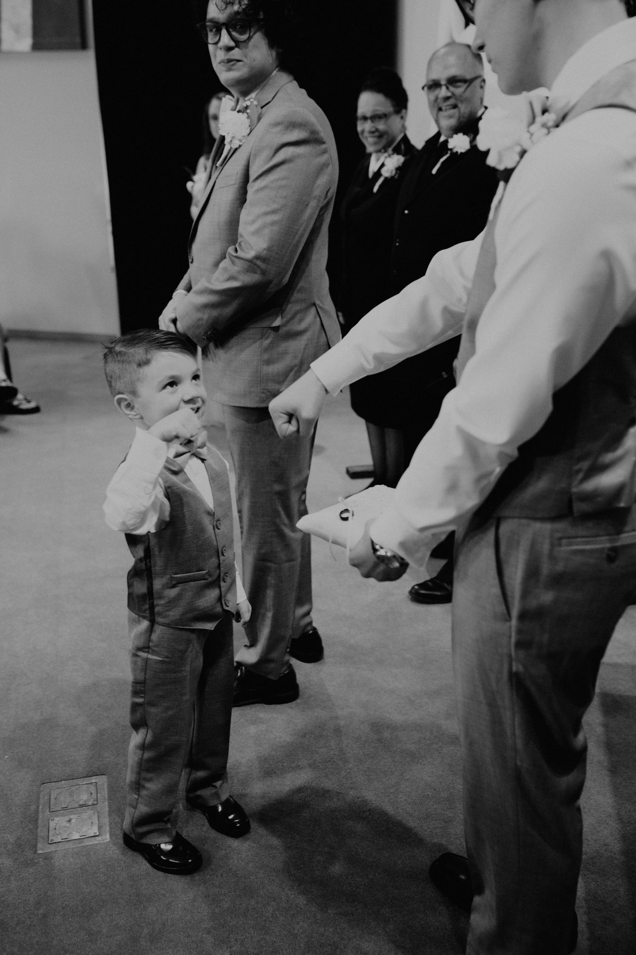 A young boy in formal attire with a vest and bow tie, giving a fist bump to a man holding an envelope, at a wedding or formal event with adults dressed in suits and dresses observing. Seattle, WA wedding photography.