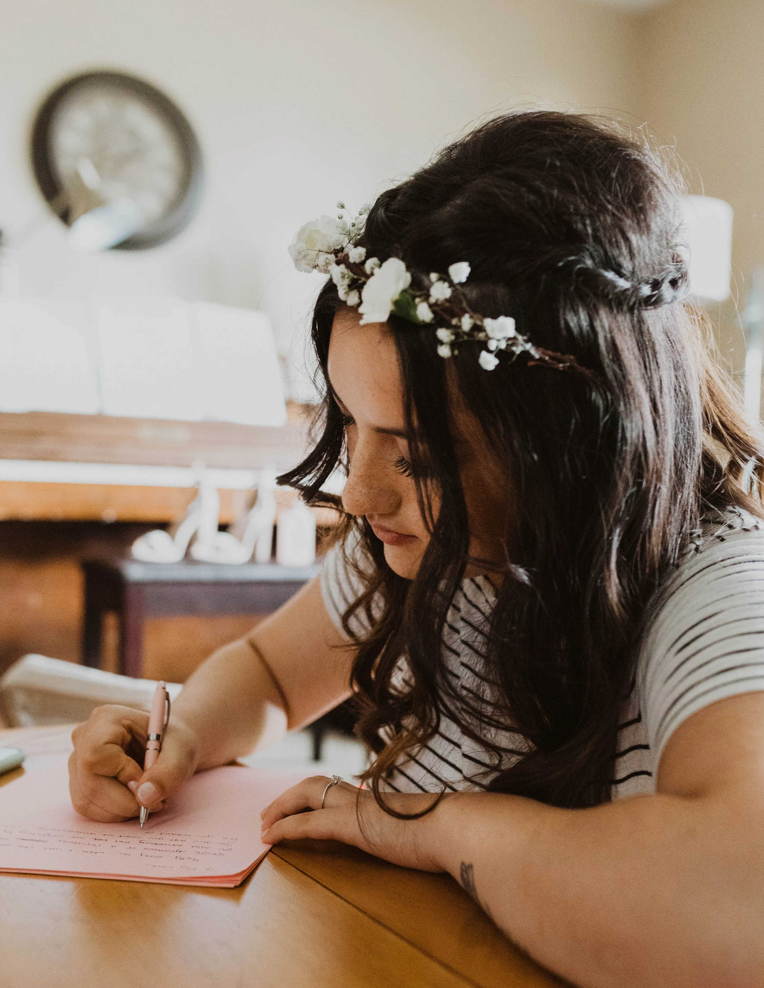 Woman with dark, wavy hair wearing a floral crown, sitting at a wooden table, writing on pink paper with a silver pen. Seattle, WA wedding photography.