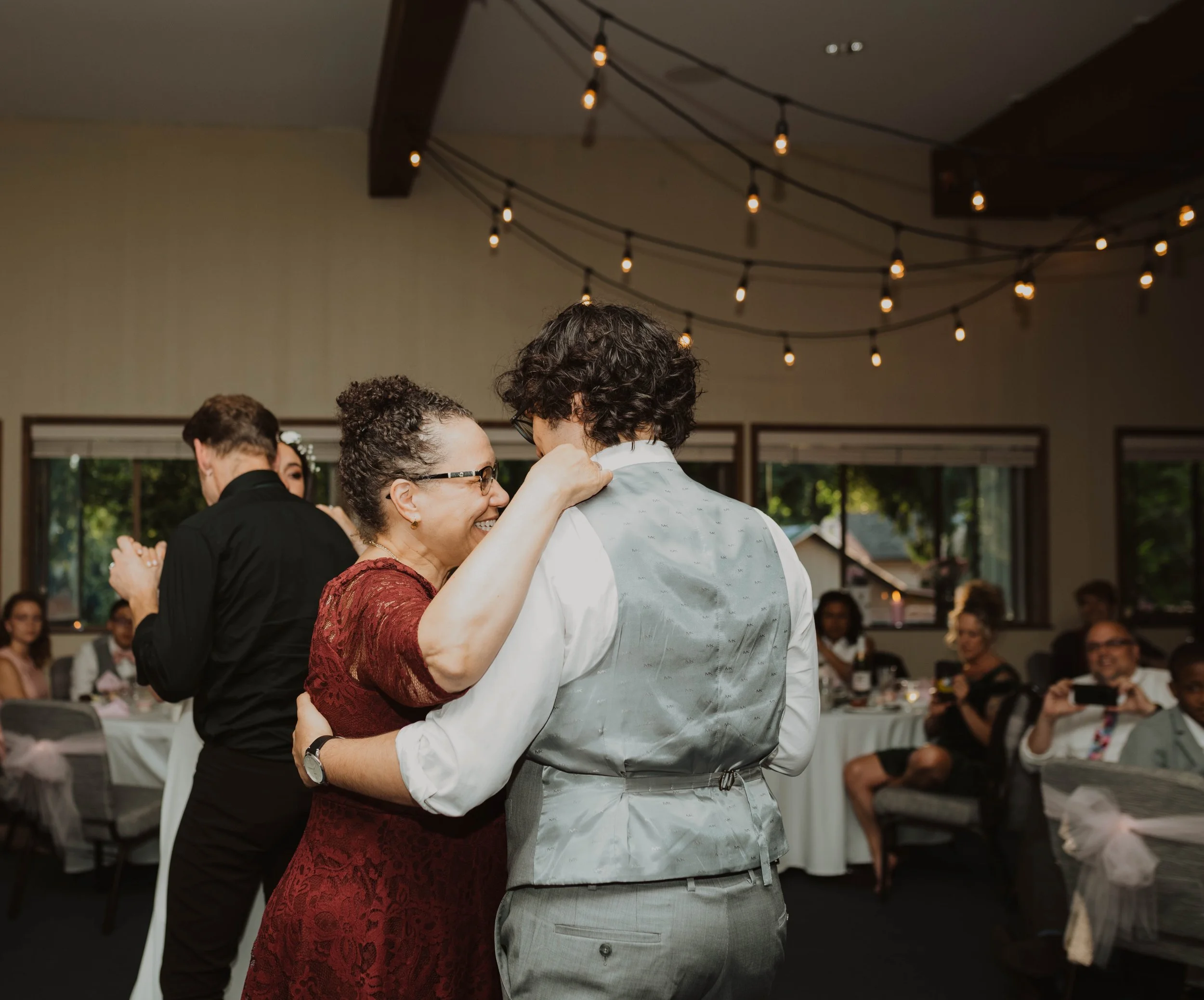 A woman in a red dress dancing with a man in a grey vest at a celebration or wedding event, surrounded by guests and decorated with string lights. Seattle, WA wedding photography.