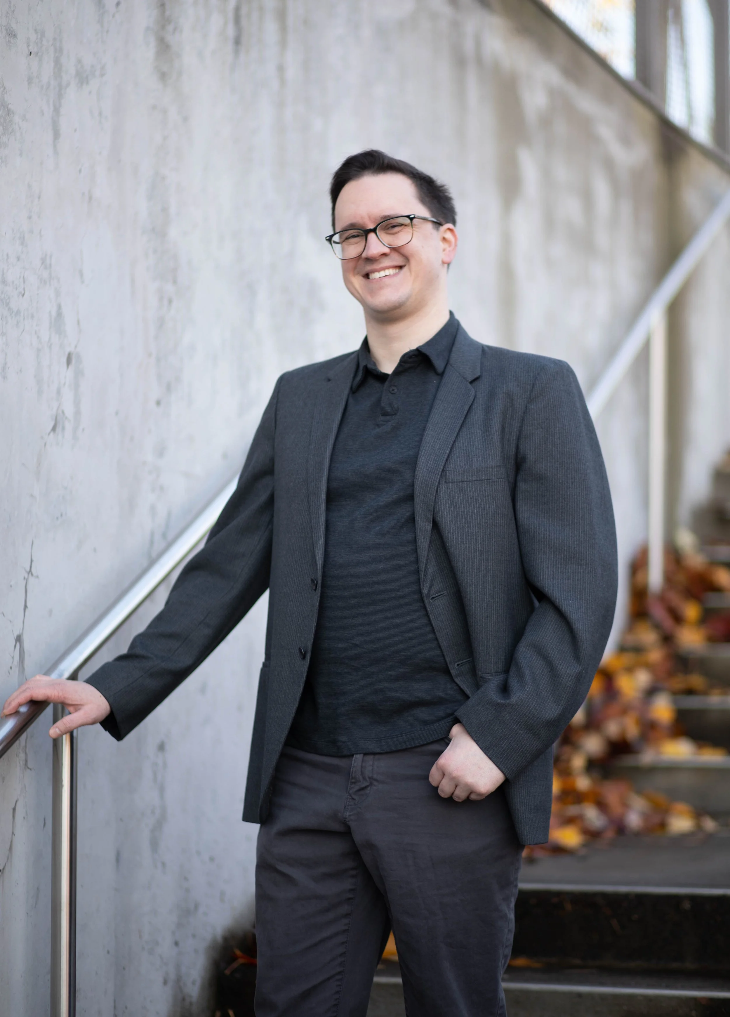 A smiling man with glasses and dark hair, dressed in a dark blazer and shirt, standing outdoors on a staircase with fallen autumn leaves around. Seattle professional head shot photography