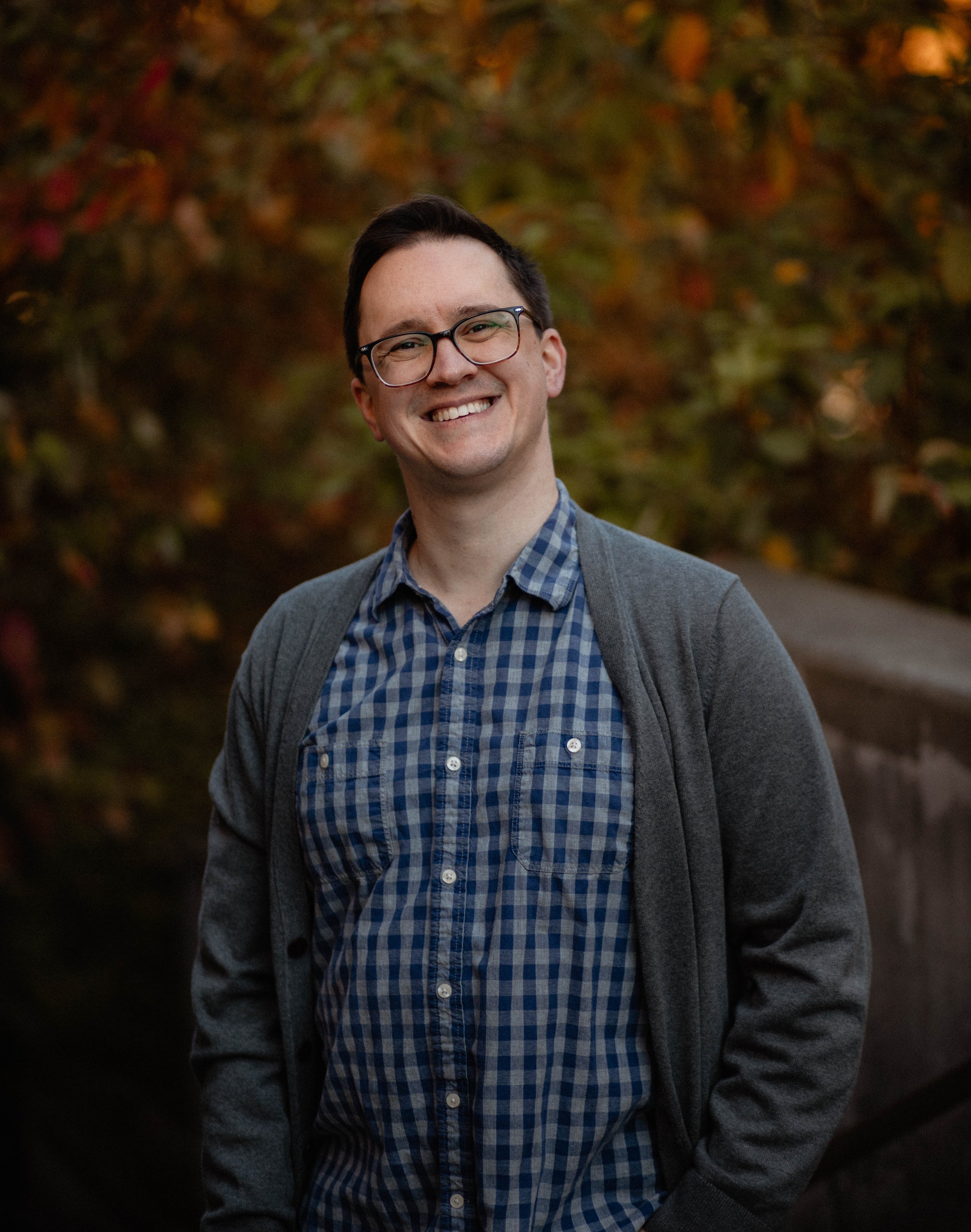 A young man with glasses, dark hair, and light skin smiling outdoors during autumn. He's wearing a blue checkered shirt, a gray cardigan, and has his hands in his pockets, standing in front of autumn foliage. Seattle professional head shot photograph
