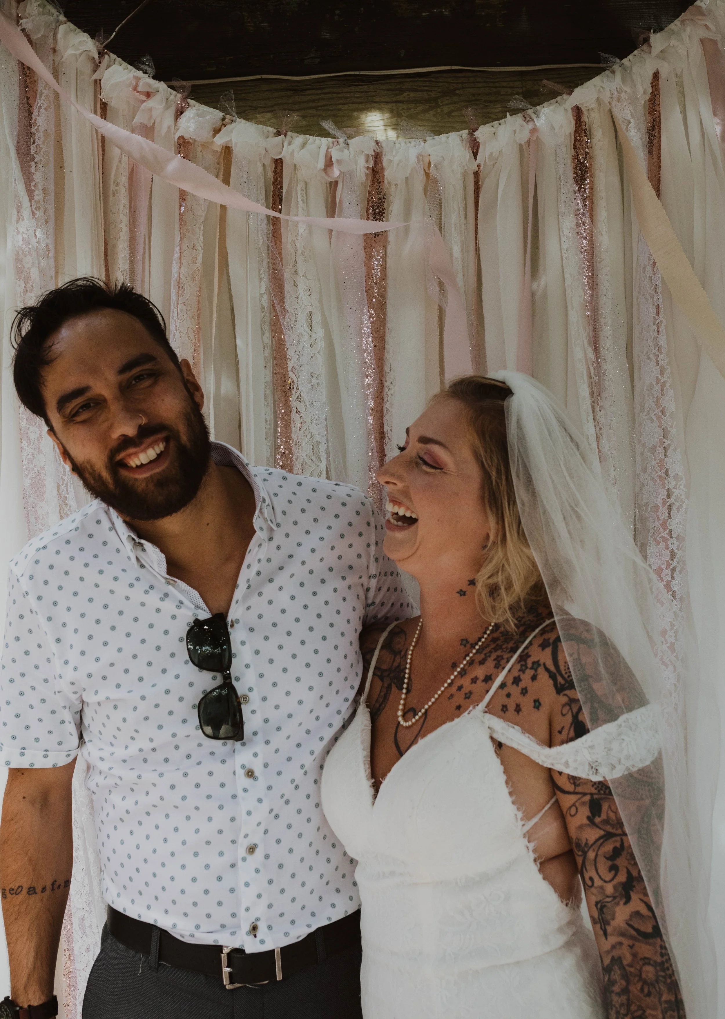 A smiling couple at a wedding, with the bride wearing a veil and tattoos on her arms and chest, and the groom wearing a white shirt with small black dots and sunglasses hanging from his shirt. They are standing in front of a pink and white fabric bac