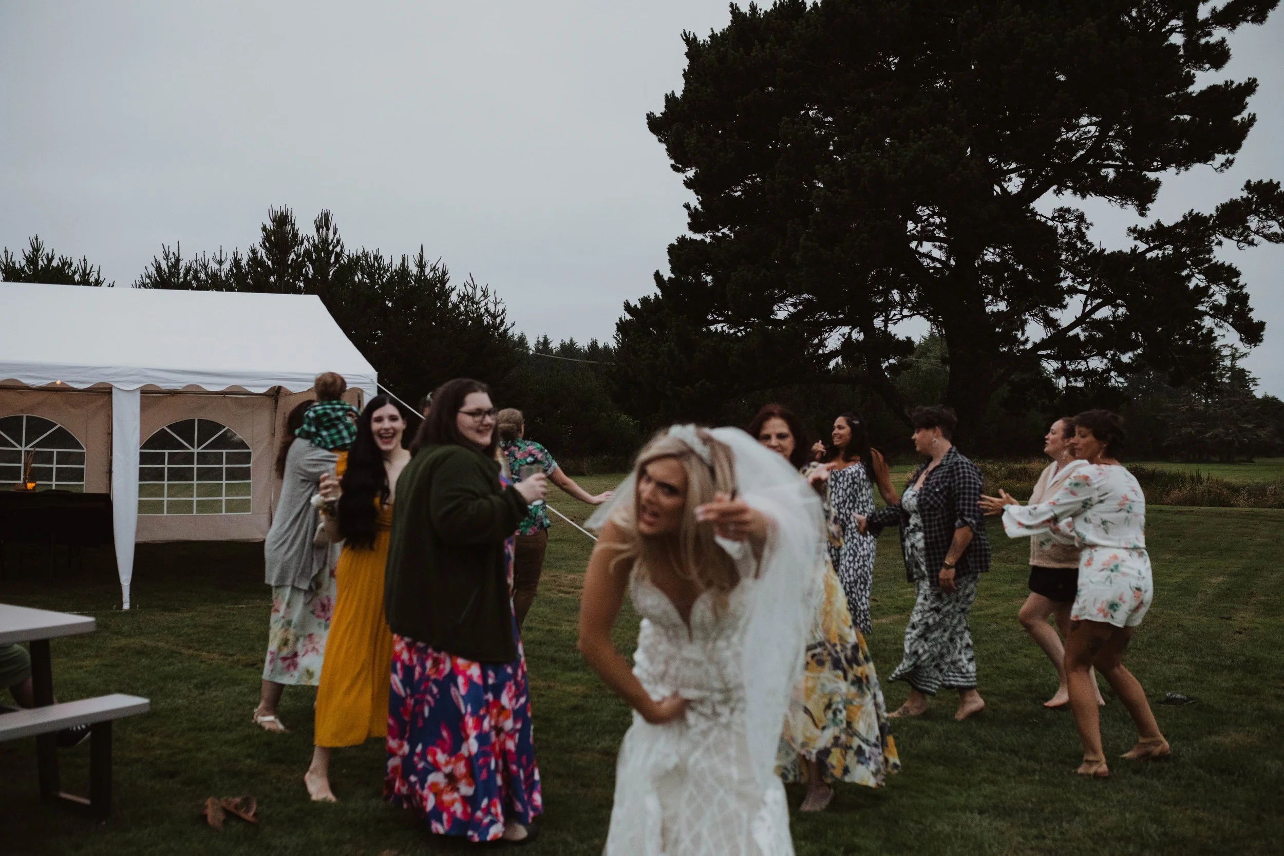 A group of women at an outdoor wedding reception dancing on the grass near a large tree, with a white tent in the background, overcast sky. Long Beach, WA wedding photography.