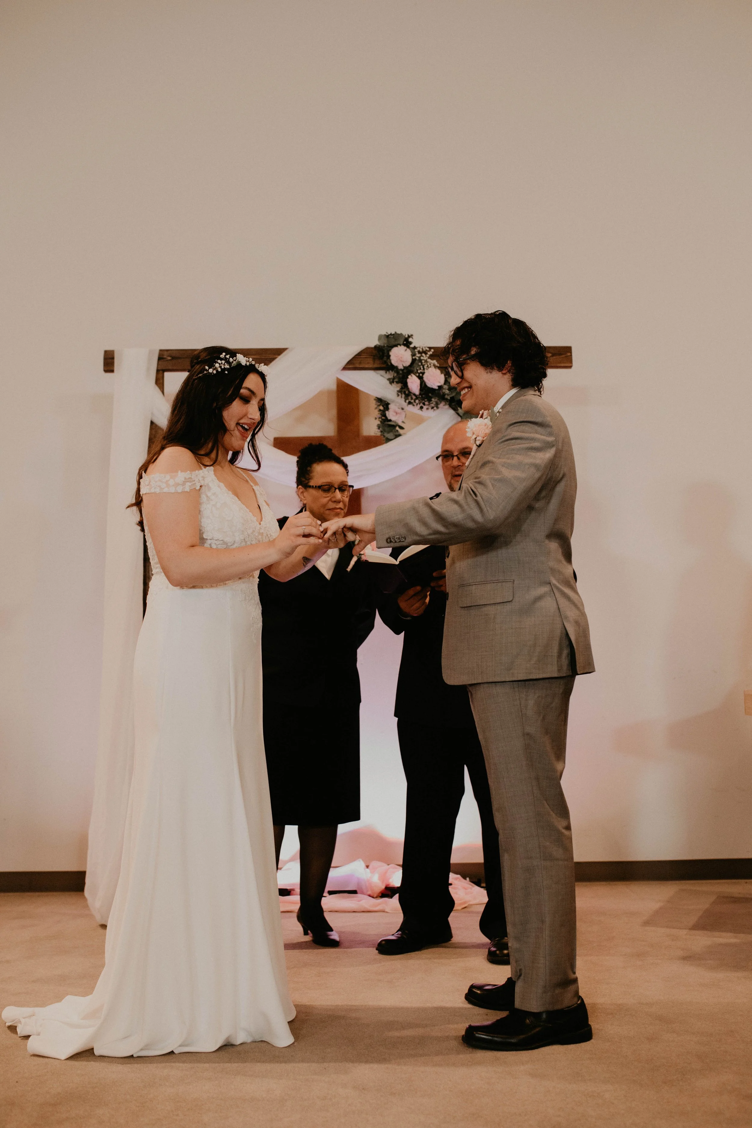 A bride and groom exchanging wedding rings during their ceremony, with officiants in the background, decorated arch with flowers in a wedding venue. Seattle, WA wedding photography.