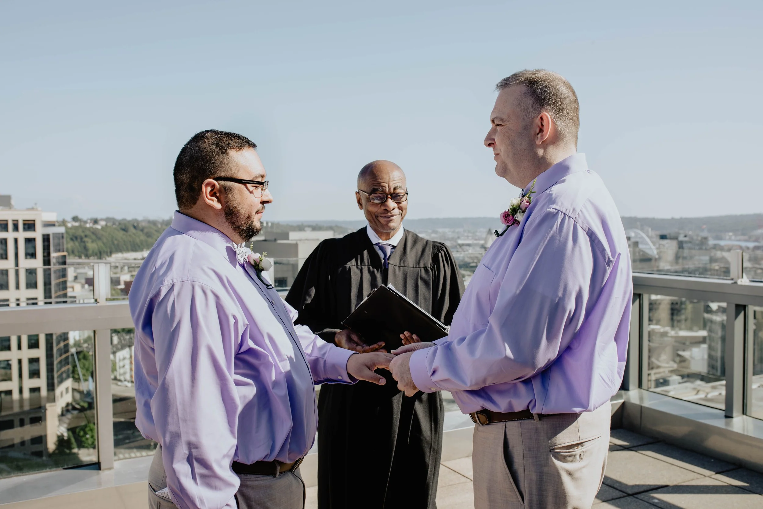 Two men in lavender shirts holding hands during a wedding ceremony with a officiant in a black robe, on a rooftop with cityscape in the background. Seattle Municipal Courthouse wedding photography.