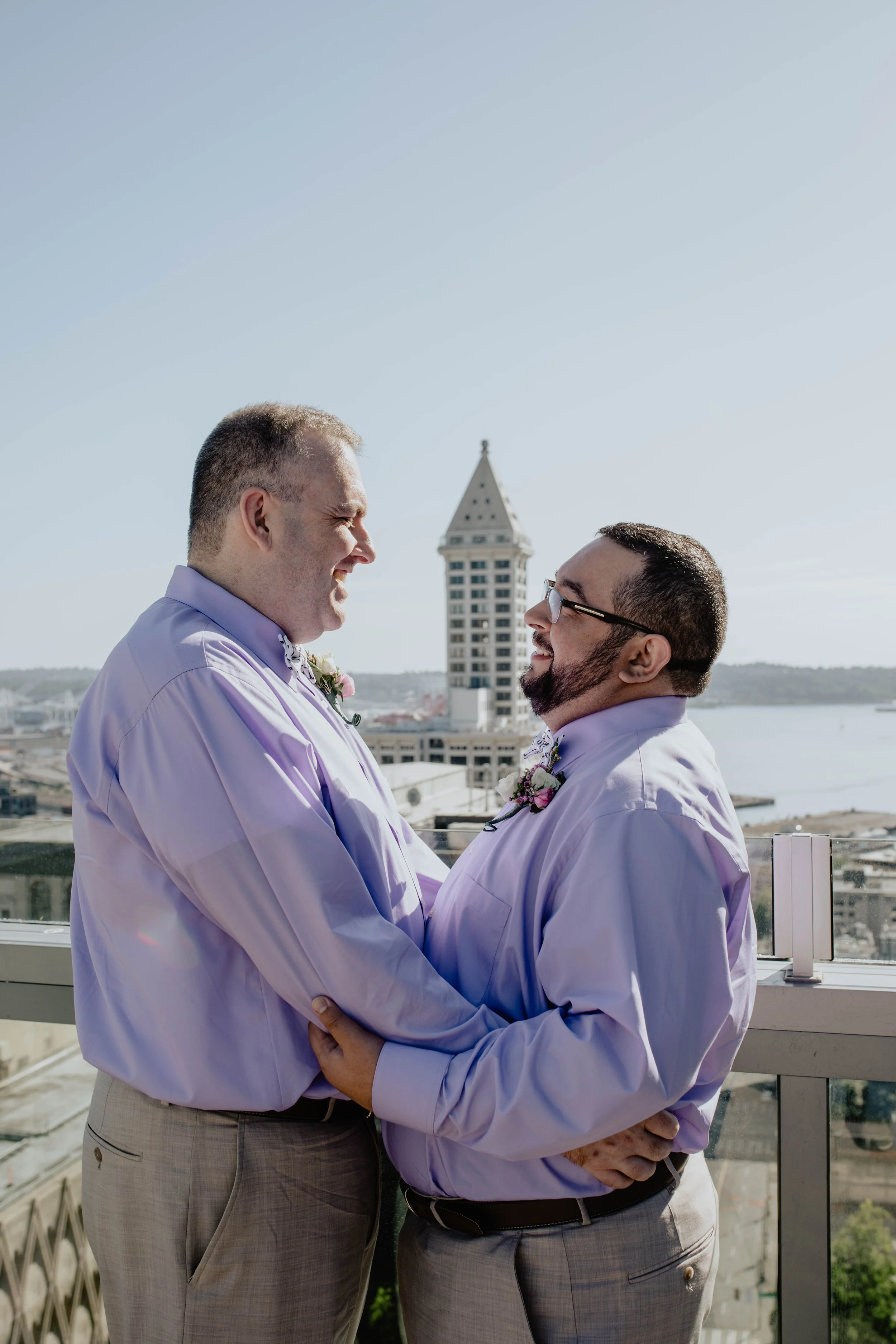 Two men in purple shirts and floral boutonnieres embrace and smile at each other on a balcony with a city skyline and water in the background. Seattle Municipal Courthouse wedding photography.