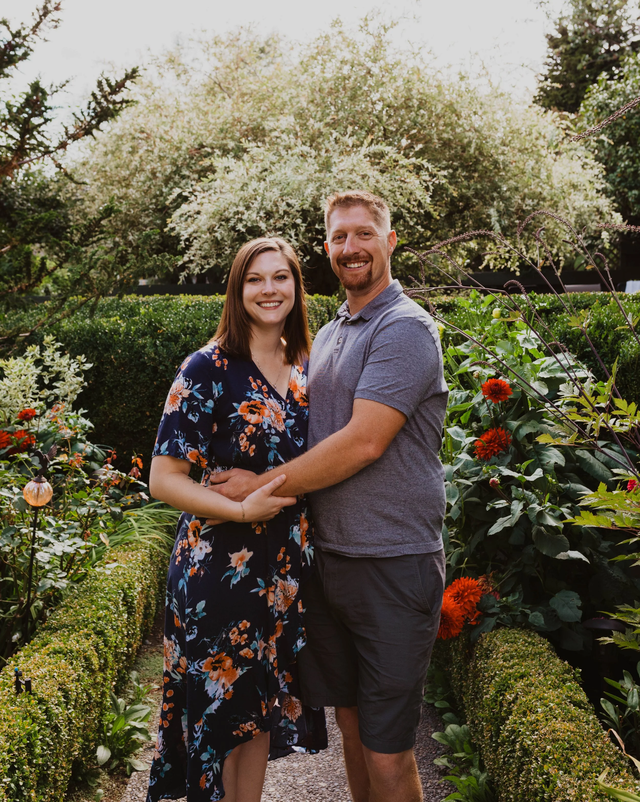 A smiling couple holding hands in a garden with vibrant flowers and lush greenery, with a large flowering tree in the background. Seattle, WA wedding photography.