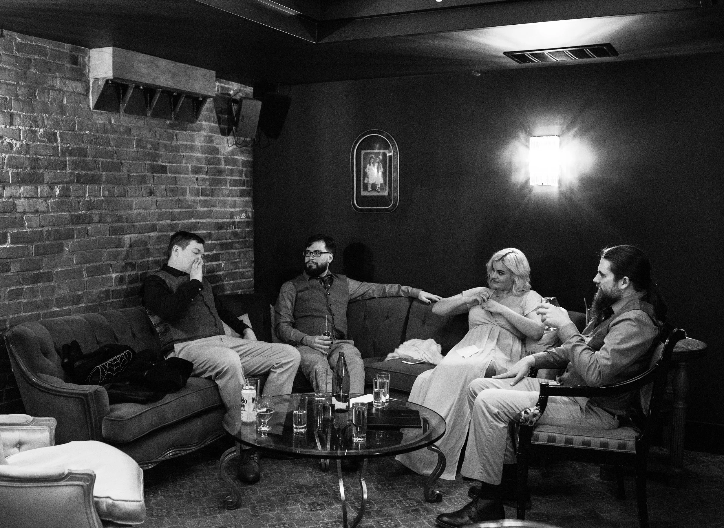 Five people sitting on a couch and chair in a dimly lit room, engaged in conversation, with drinks on a small table in front of them. Pioneer Square, Seattle, WA wedding photography.