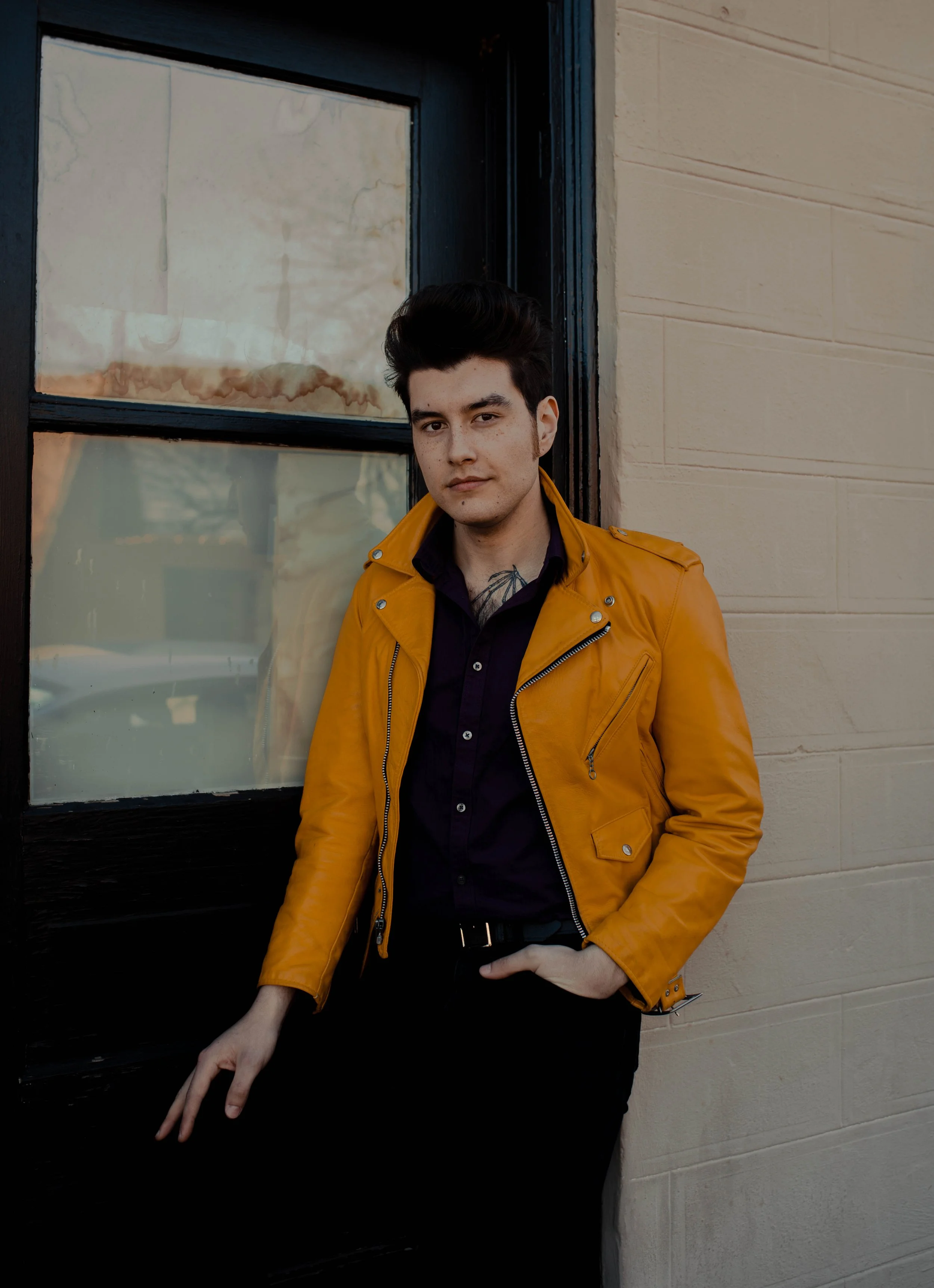 A young man with dark hair and tattoos, wearing a yellow leather jacket over a black shirt, standing against a black window frame and beige wall. Seattle professional head shot photography