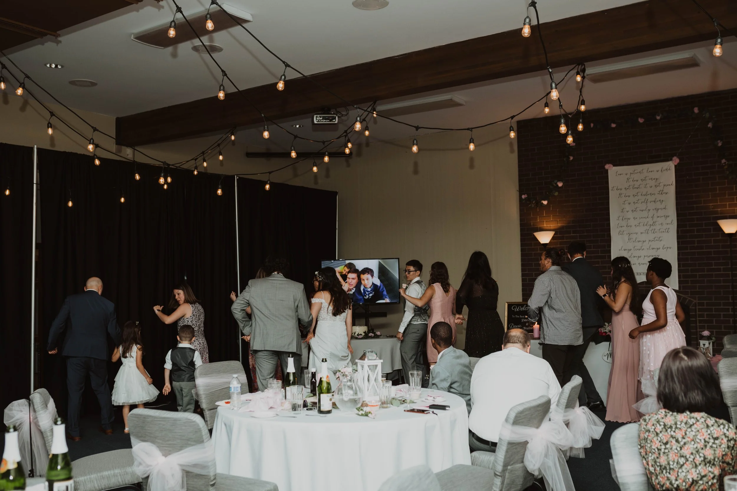 People dancing and socializing at a celebration in a decorated banquet hall with string lights, tables with bottles and glasses, and a large screen showing children. Seattle, WA wedding photography.