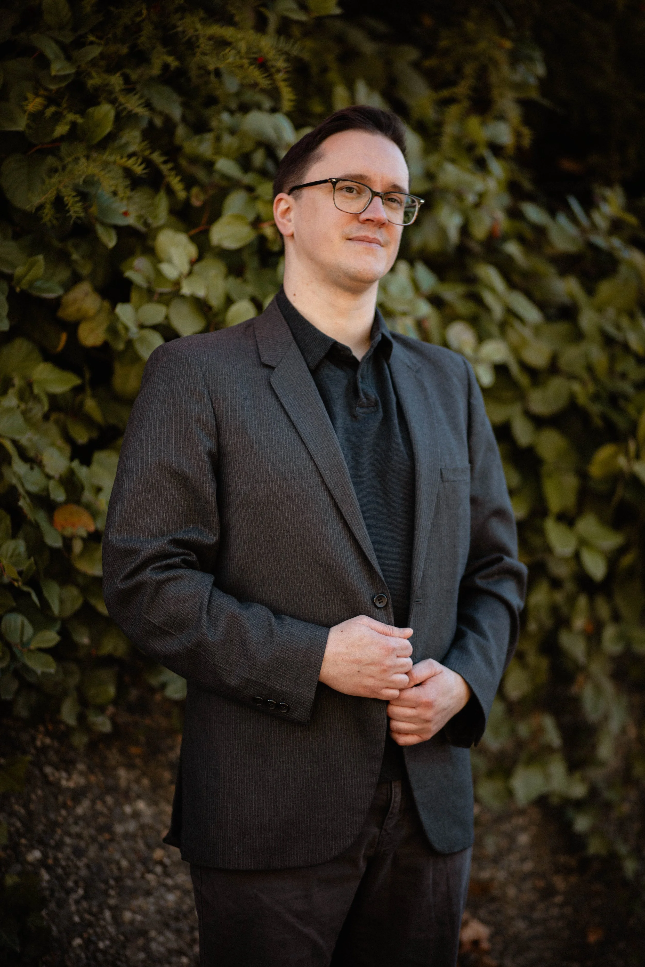 A man wearing glasses in a dark suit jacket standing outdoors in front of a leafy background Seattle professional head shot photography