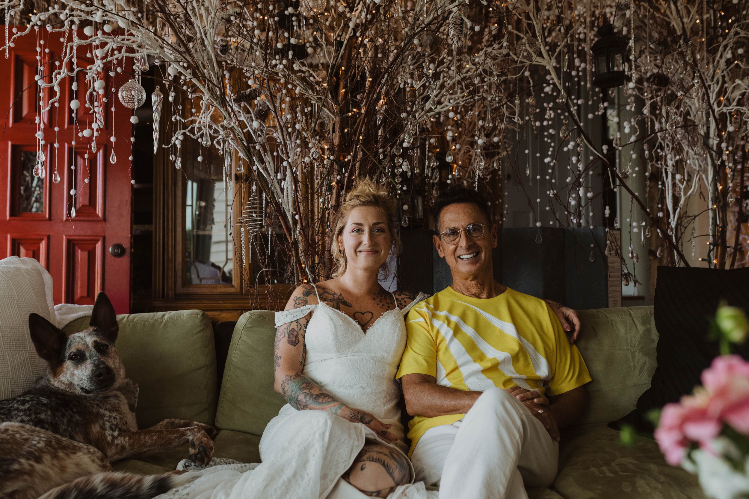 Two people sitting on a sofa in a decorated room with a dog lying beside them. The background features branches with hanging ornaments and string lights. Seattle, WA wedding photography.