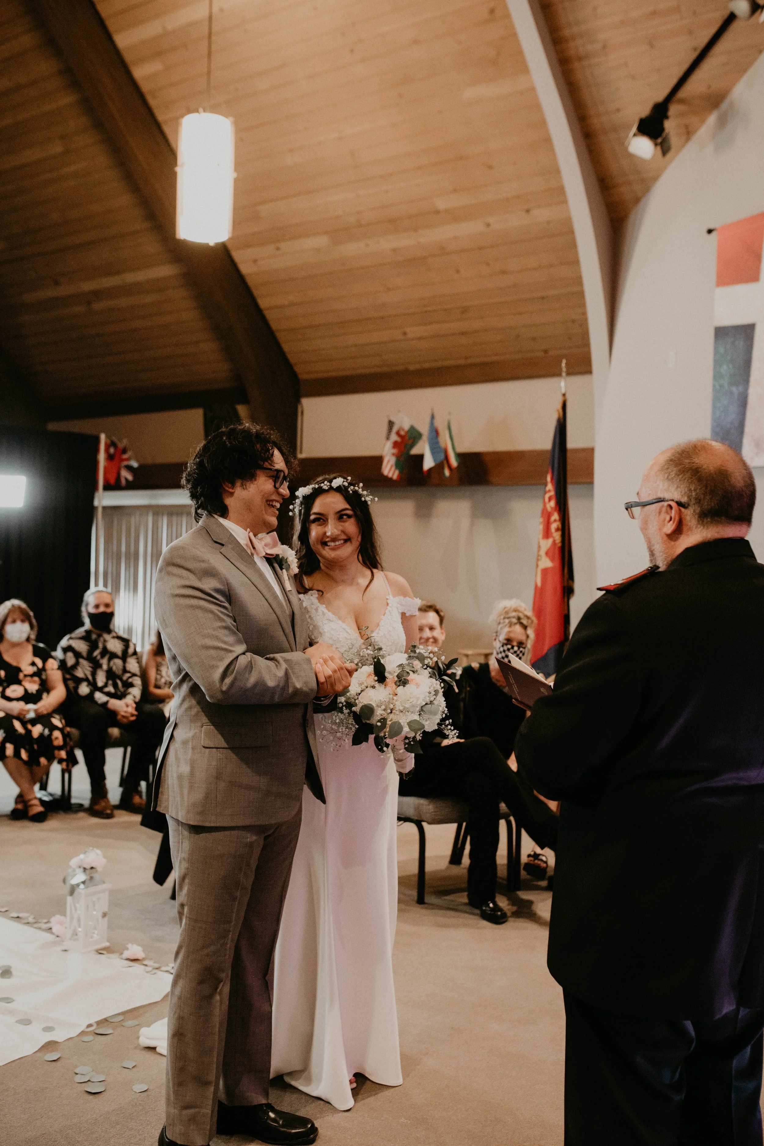 A wedding ceremony with a couple standing before an officiant. The bride is holding a bouquet and wearing a white wedding gown with off-shoulder sleeves. The groom is wearing a light gray suit. Guests are seated in the background, some wearing masks,