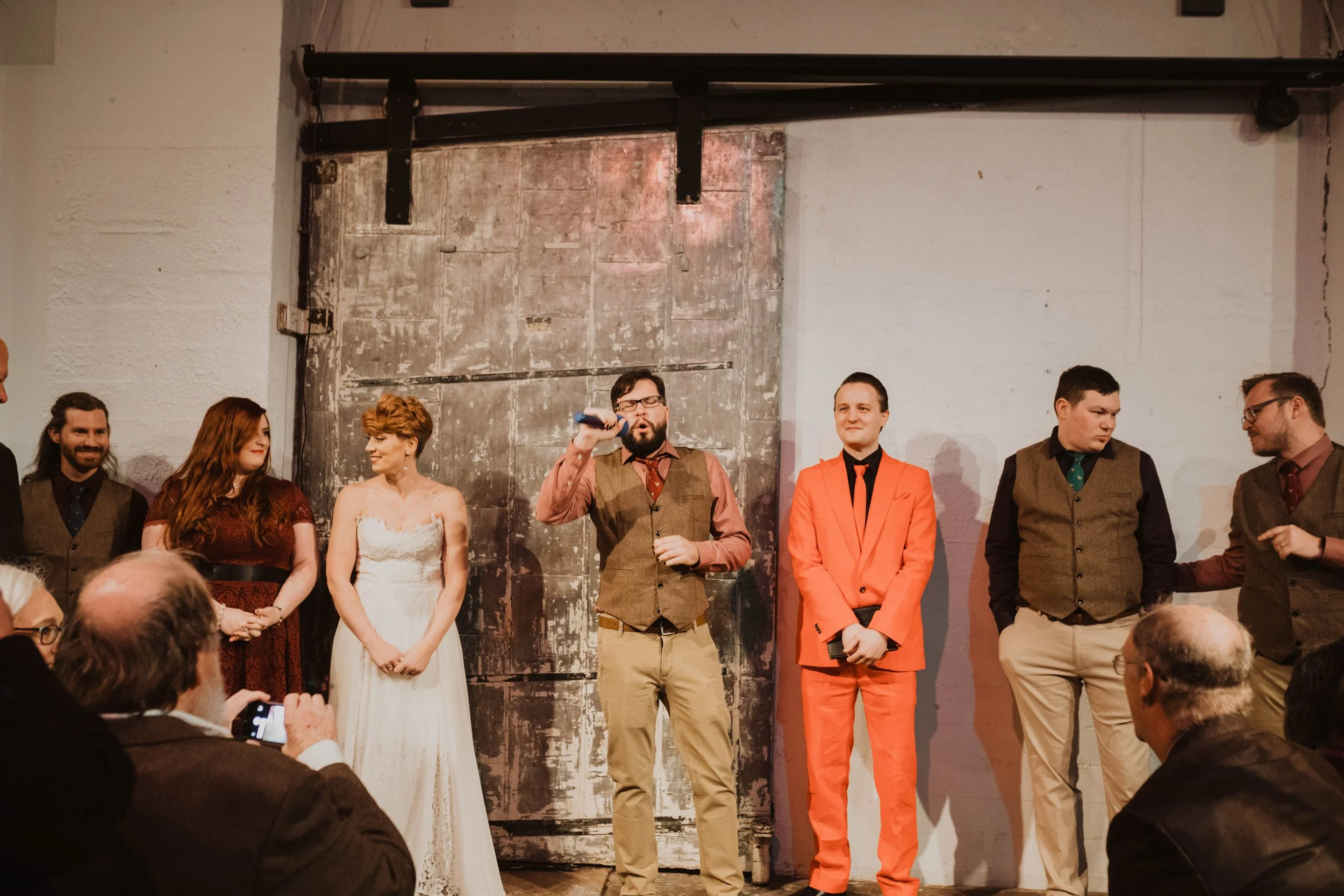 Group of people on stage at a formal event. One man is speaking into a microphone while others stand nearby, including a woman in a white dress and a man in a bright orange suit. Pioneer Square, Seattle, WA wedding photography.
