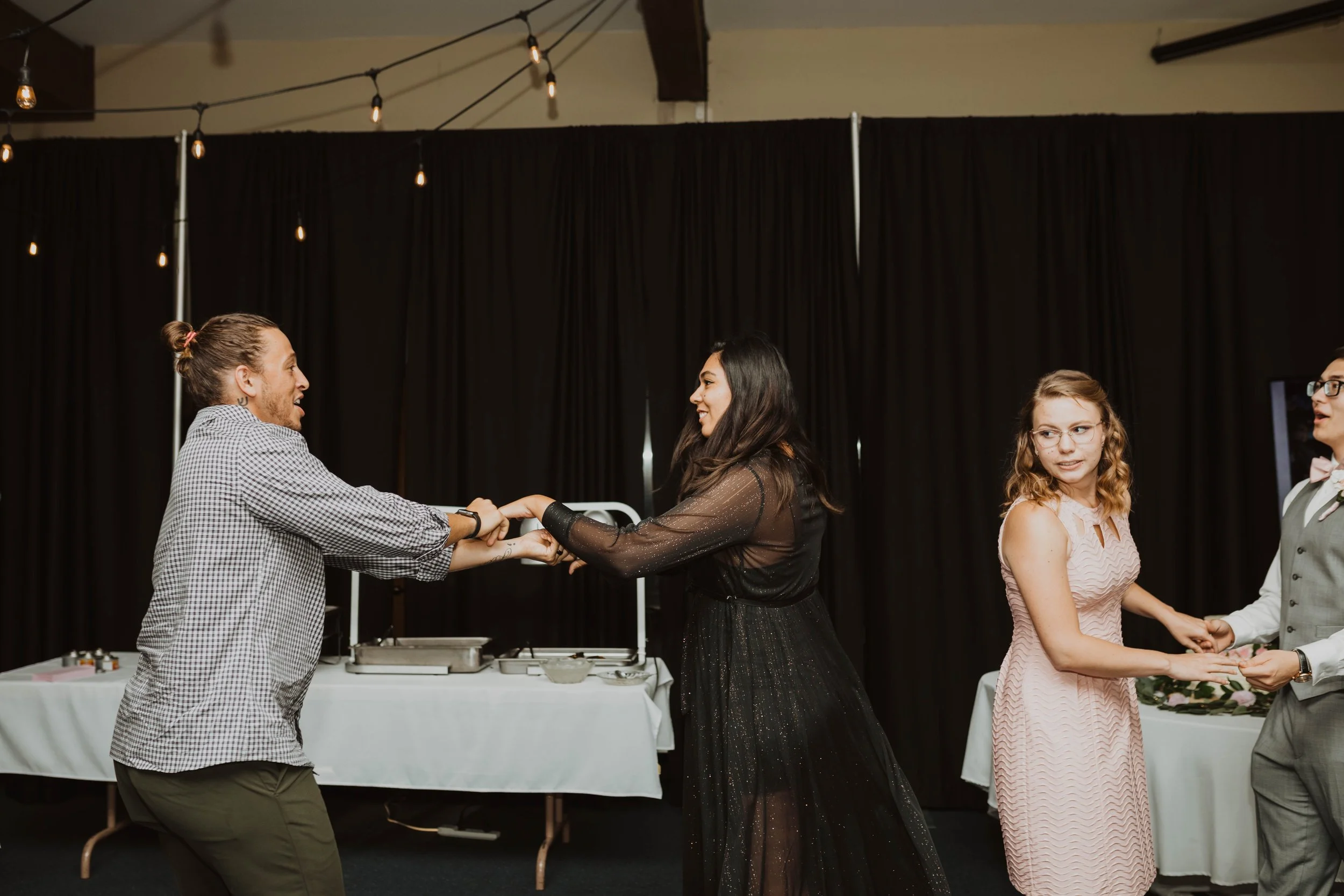 Two couples dancing at a wedding reception with string lights overhead and black curtains in the background. Seattle, WA wedding photography.