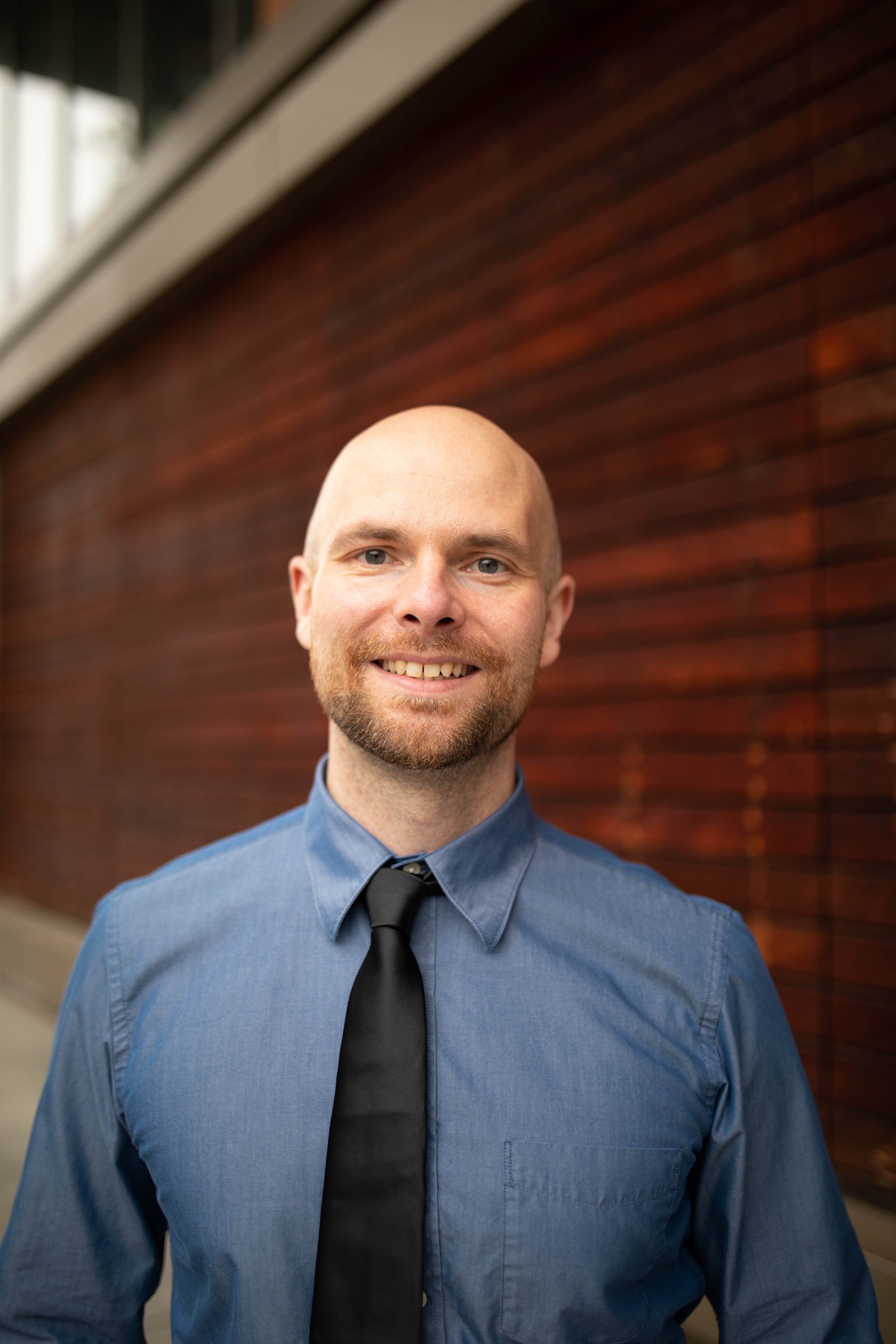 Portrait of a smiling man with a beard, wearing a blue dress shirt and black tie, standing outdoors in front of a wooden wall. Seattle professional head shot photography